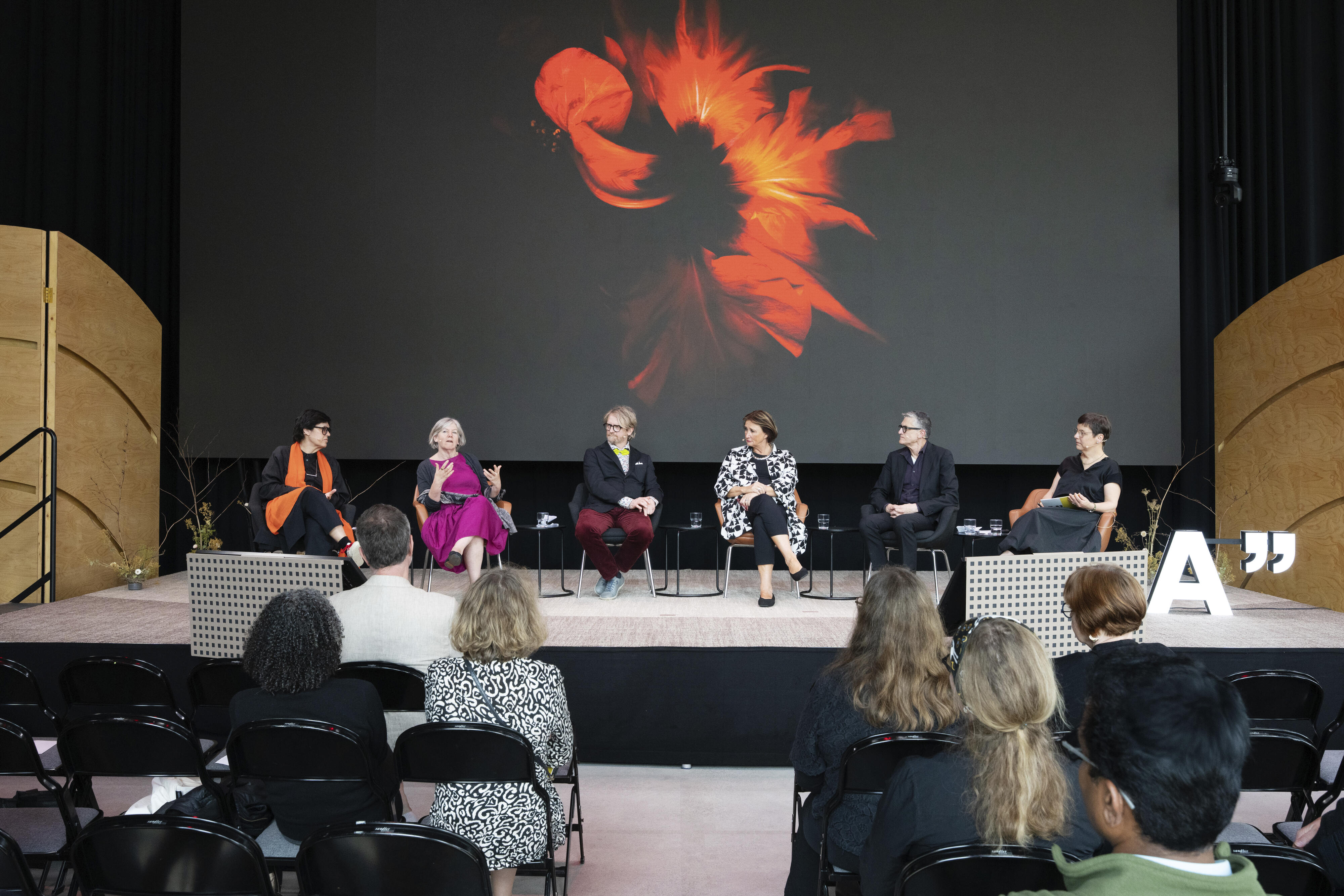 Six people sit on stage discussing, behind them a large screen showing abstract floral art. Audience watches attentively.