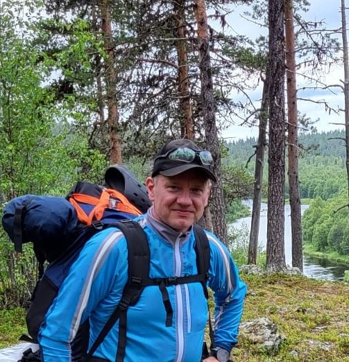 Siniseen takkiin ja lippalakkiin pukeutunut henkilö vaeltaa ison repun kanssa. Taustalla näkyy puita ja joki.