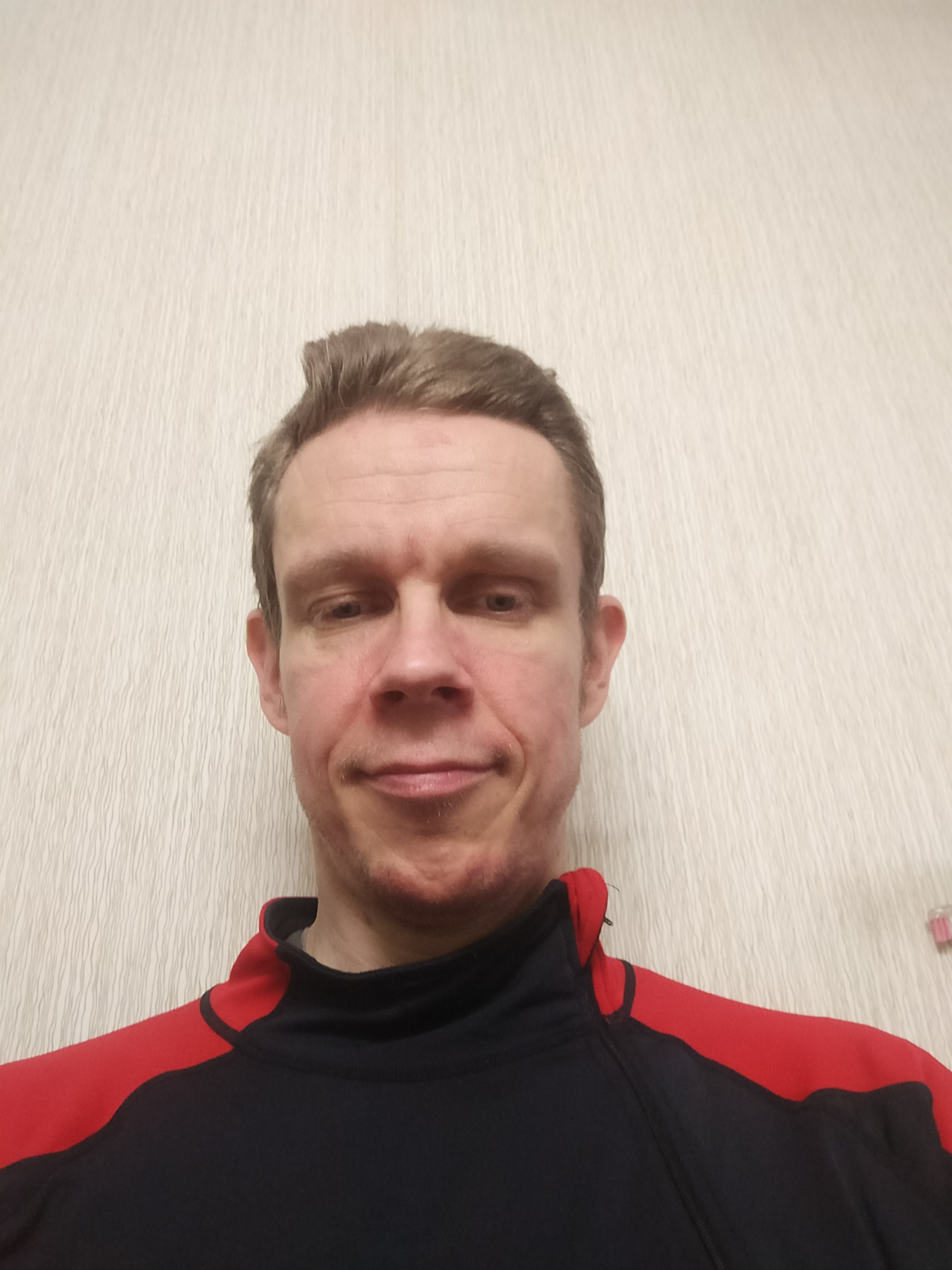 Henkilö, jolla on lyhyet ruskeat hiukset ja päällään musta ja punainen paita, seisoo beigellä taustalla.