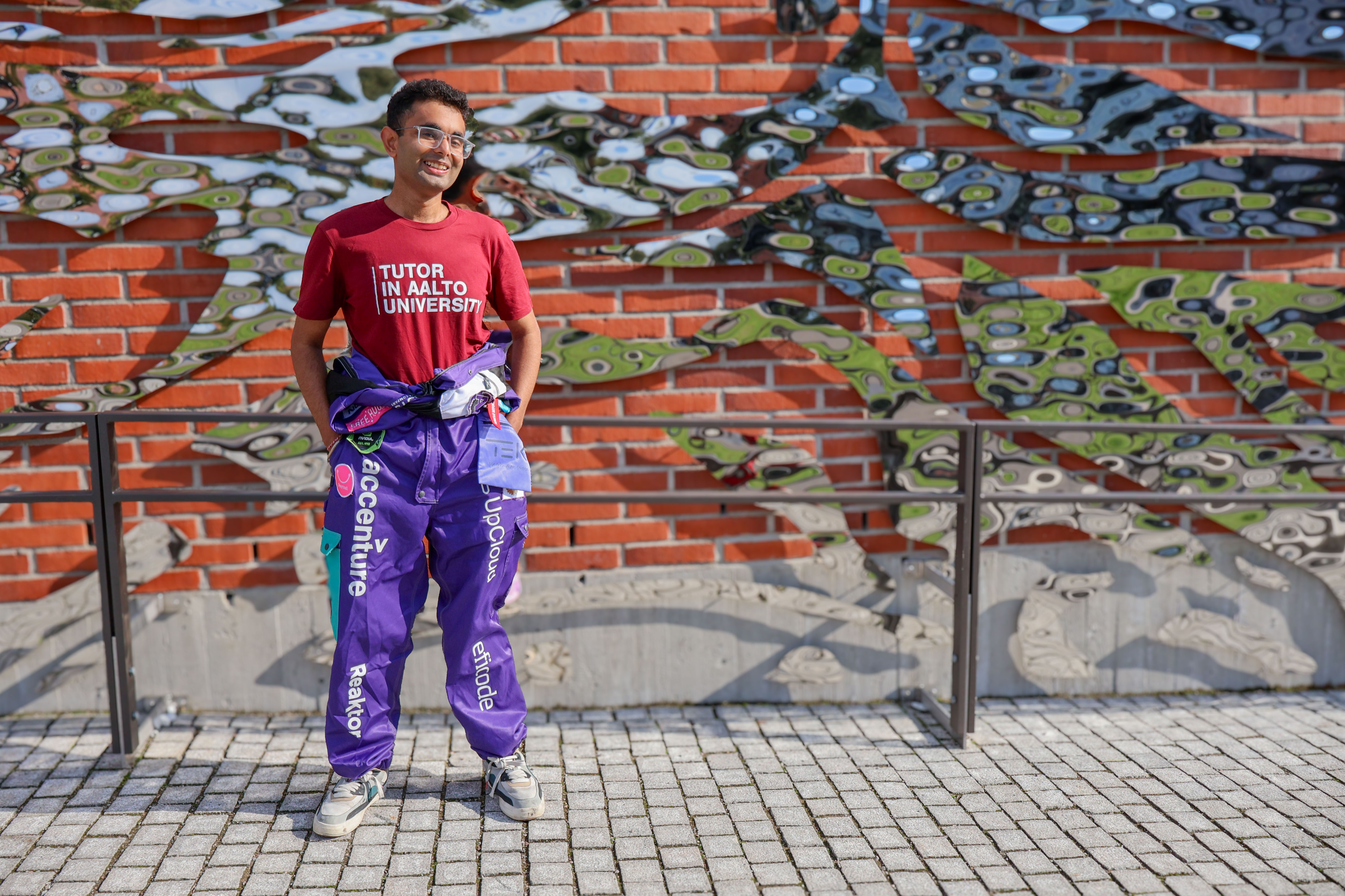 Hitesh Monga wearing Tutor in Aalto University shirt and overalls, standing in front of a brick wall with metal artwork