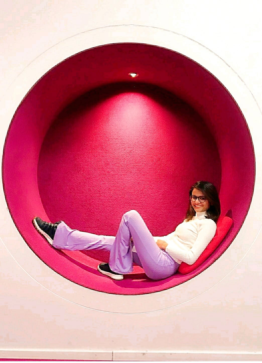 Person in white jumper and lilac trousers reclines in round red alcove with spotlight above.