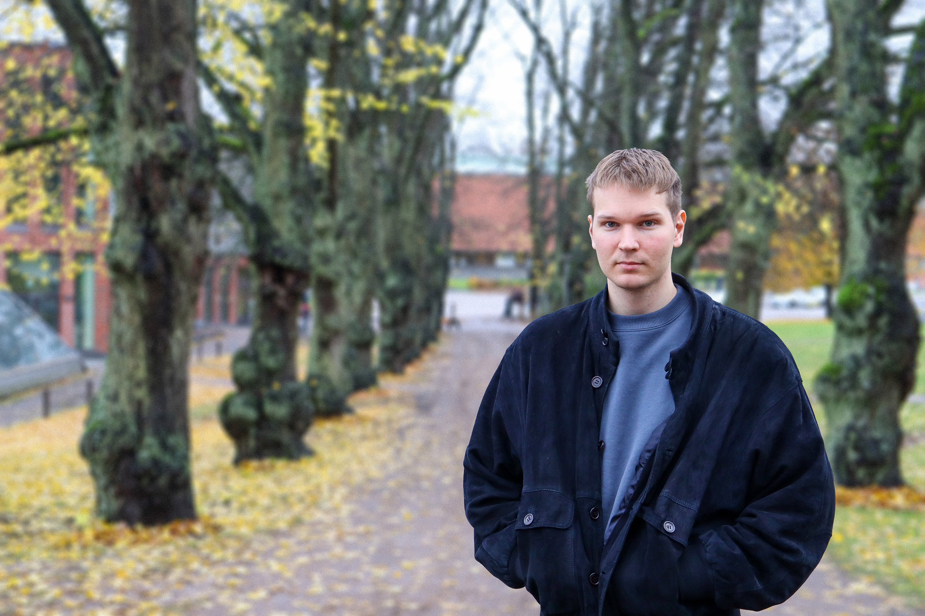 Henkilö seisoo polulla syksyisten puiden ympäröimänä, joissa on keltaisia lehtiä. Hänellä on tumma takki ja harmaa paita.