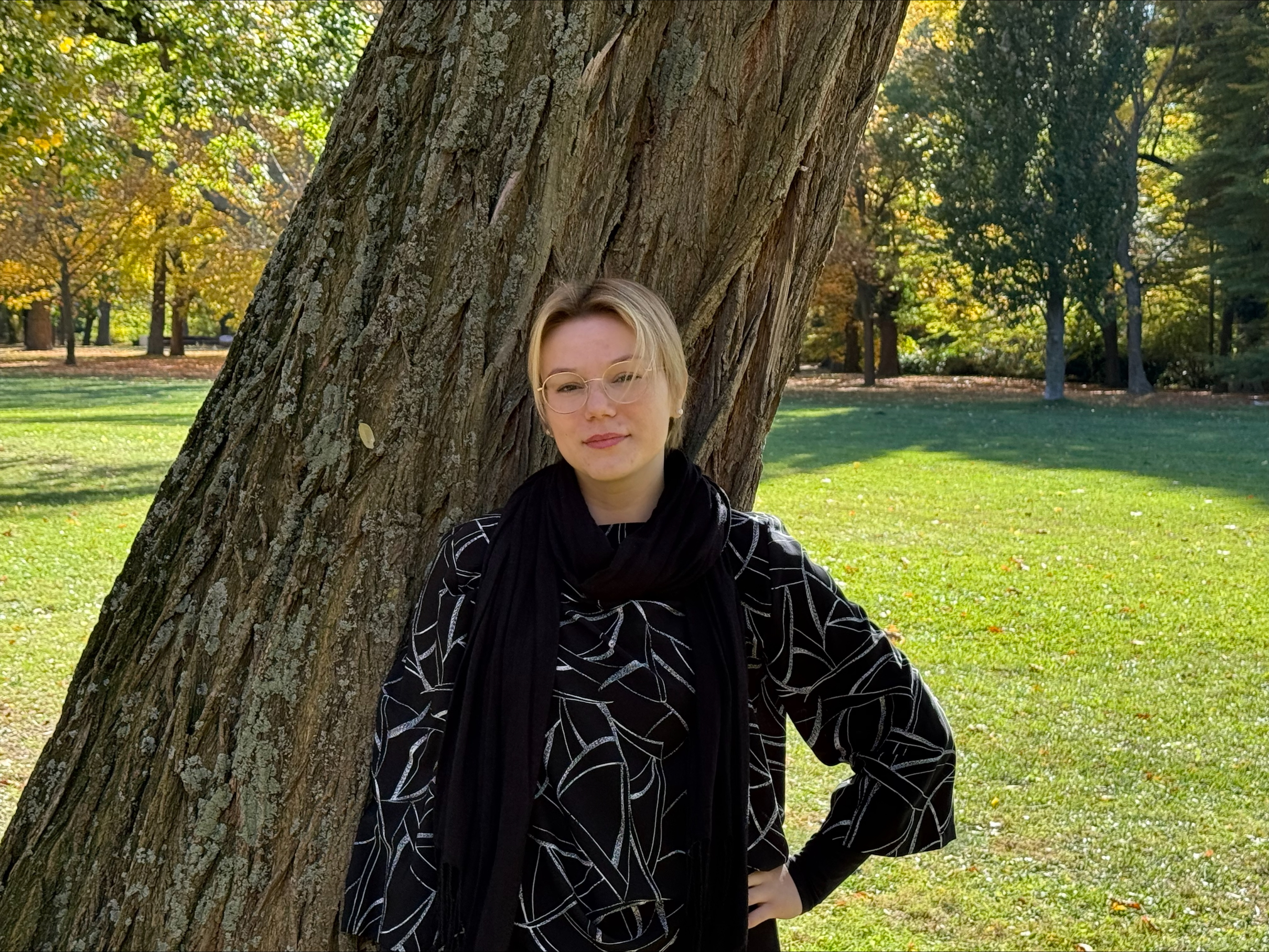 Person with hand on hip leans against large, textured tree trunk in a sunlit park with autumn foliage.