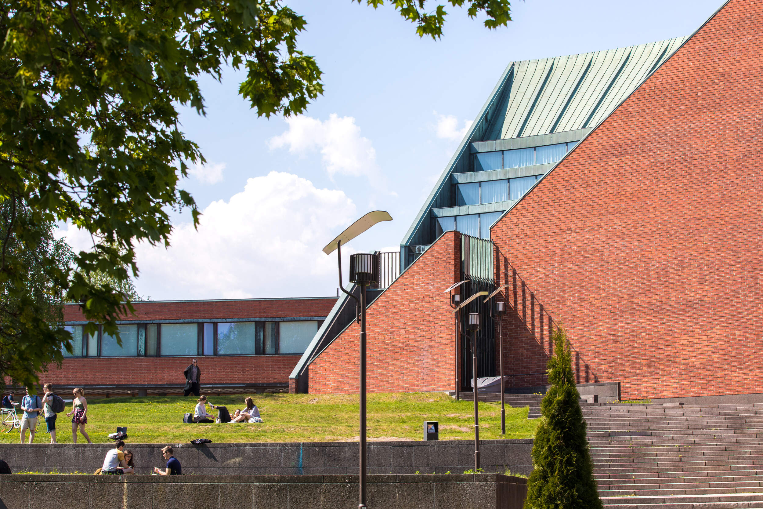 People relax on a grassy area near a modern building with red brick walls and large glass windows under a blue sky.