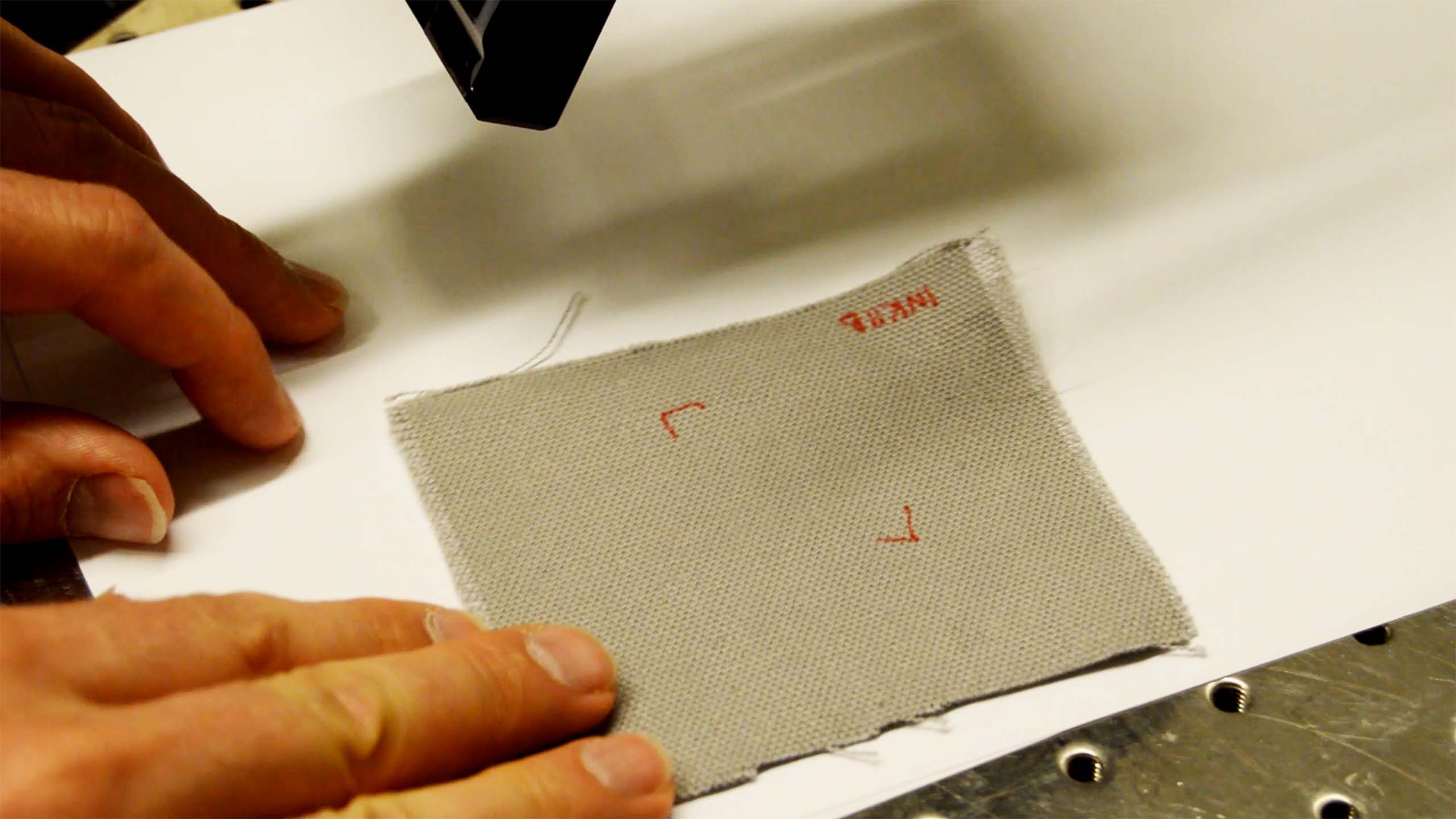 Hands positioning a grey fabric swatch with red markings under a machine on a white surface.