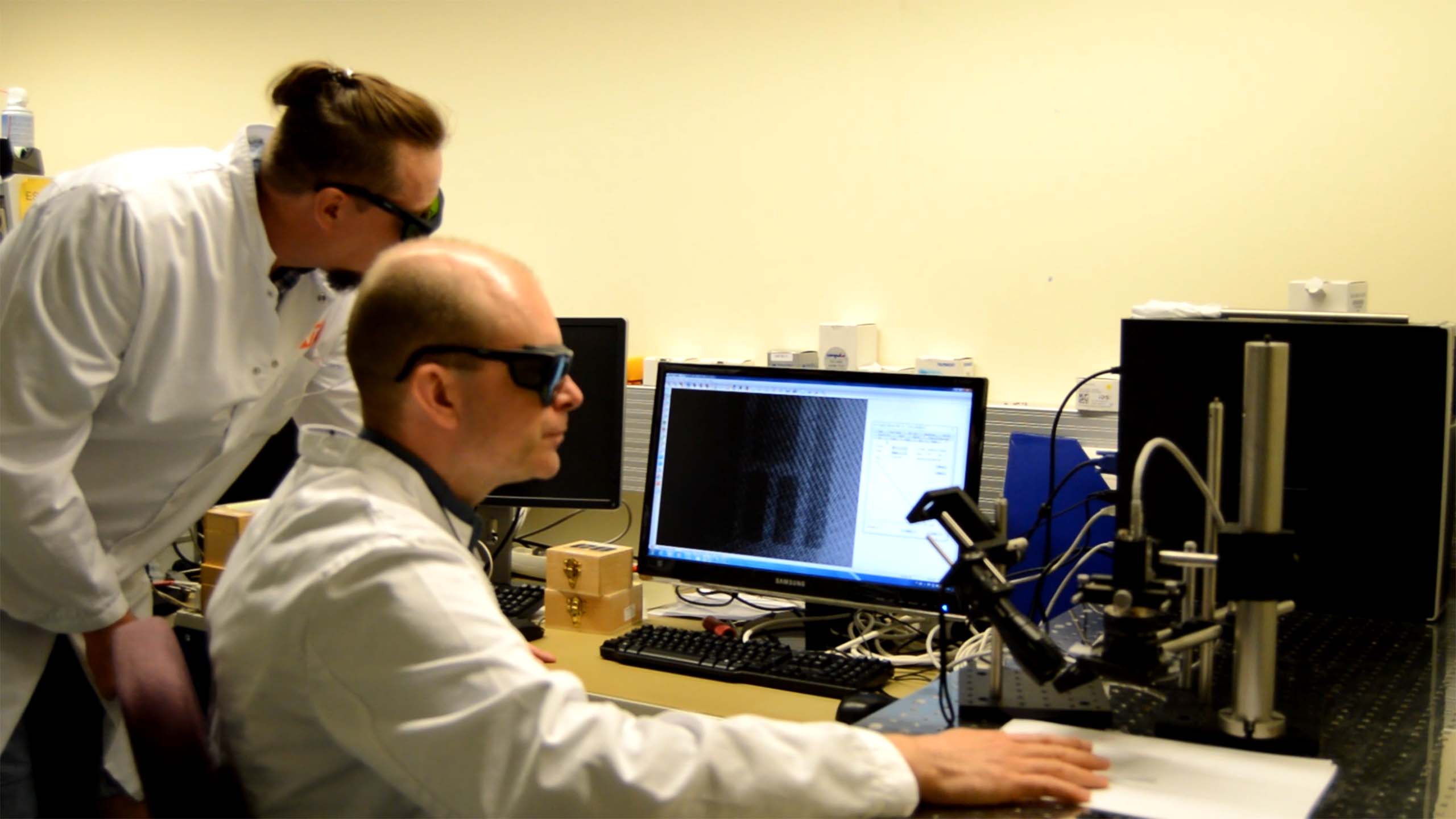 Two scientists in lab coats working at a computer with scientific equipment on the desk.
