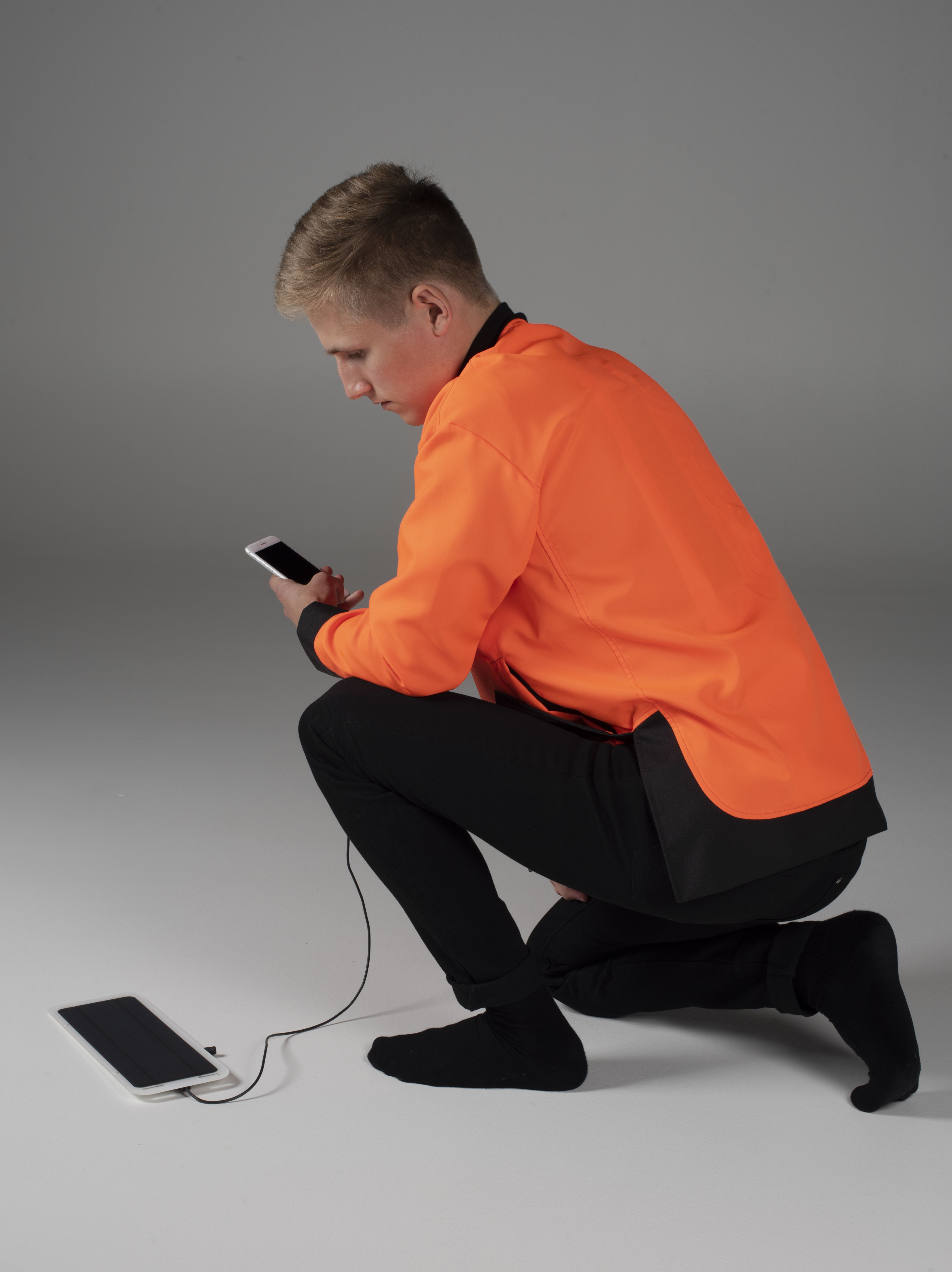 Person in an orange jacket kneeling and using a smartphone connected to a tablet on the floor.