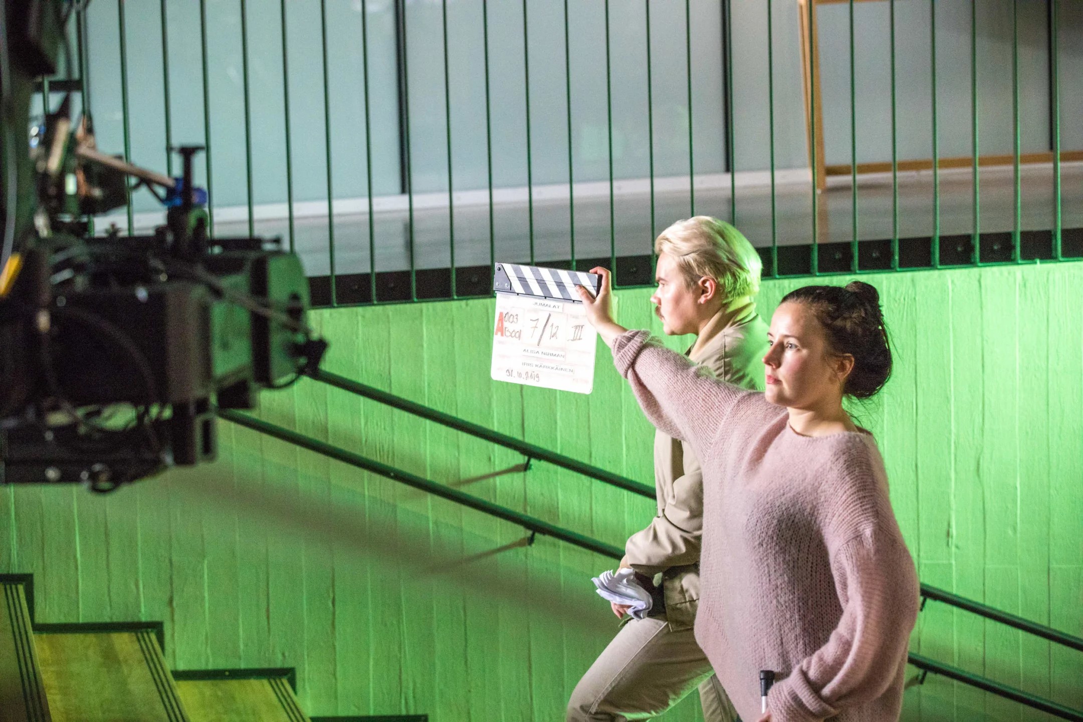 Two people on a film set, one holding a clapperboard. A camera rig is visible on the left. Green wall in the background.