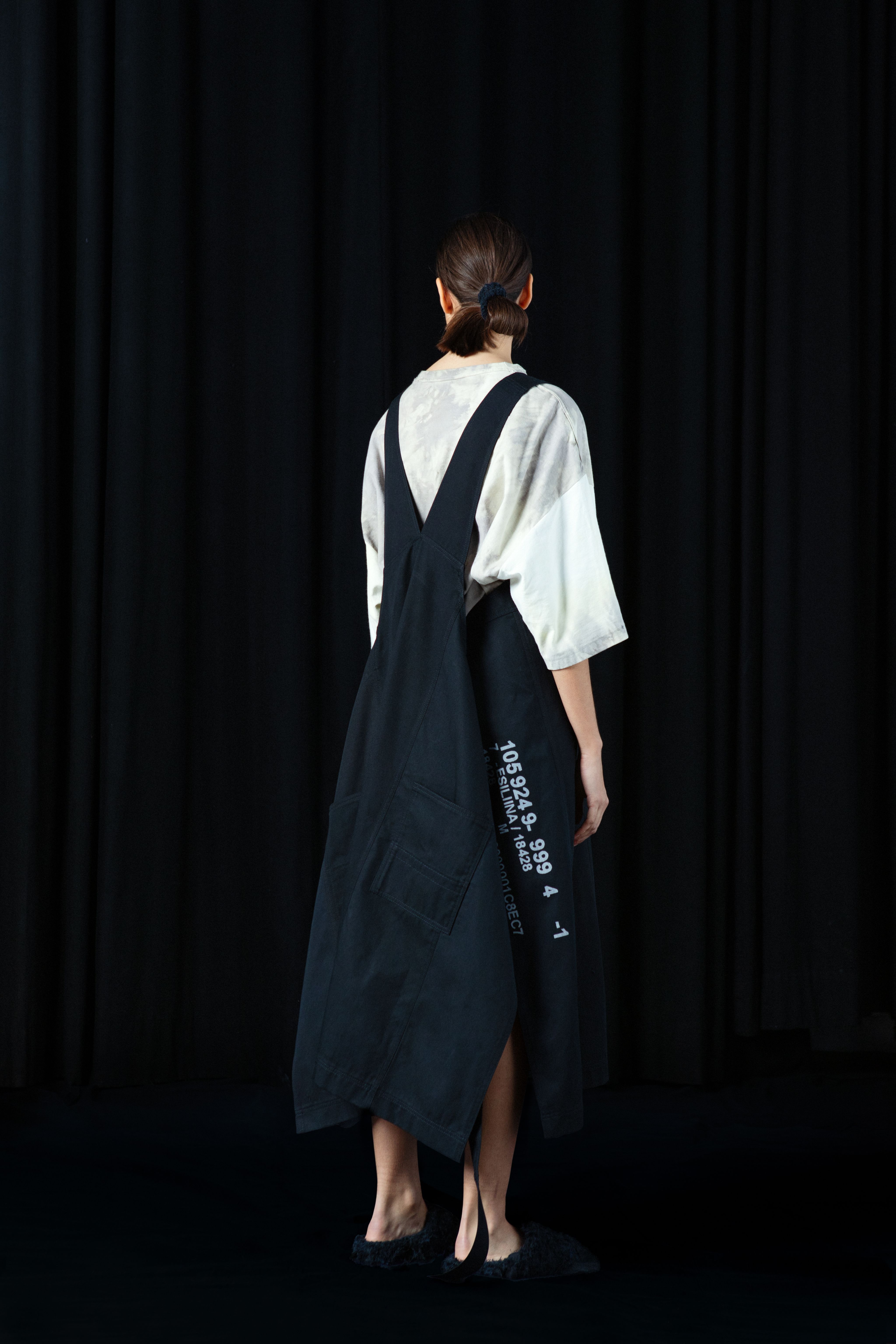 A person with a ponytail wearing a white shirt and black apron stands facing away in front of a dark curtain.