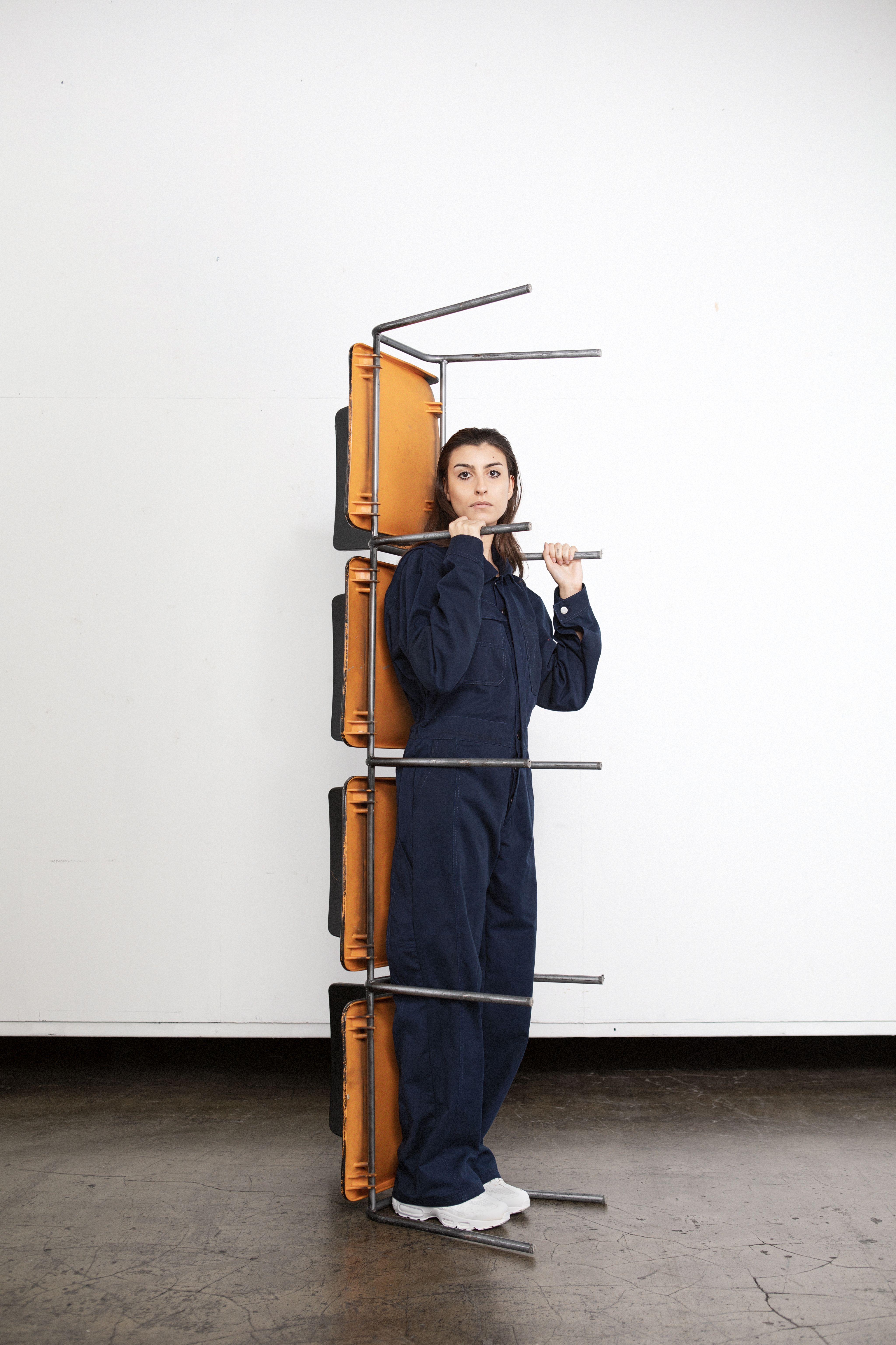 Person in blue overalls standing behind a stack of orange chairs held by metal bars.