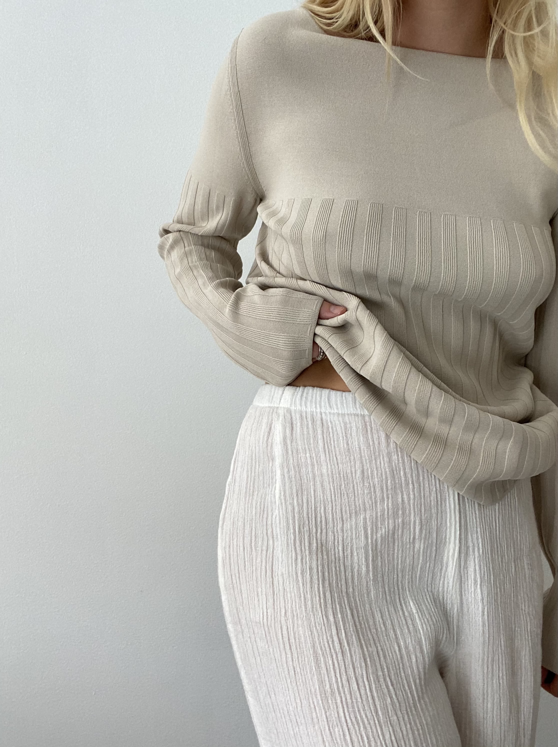 A person wearing a beige ribbed jumper and white textured trousers, partially tucking the jumper into the trousers.