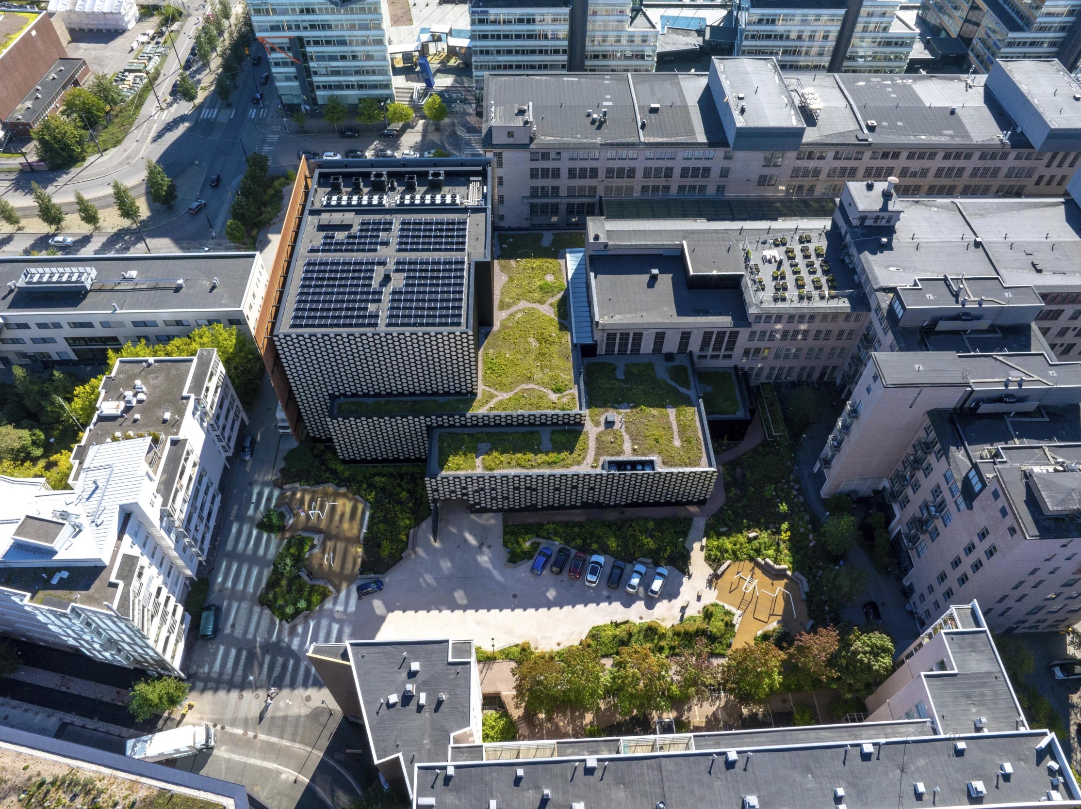 Aerial view of urban buildings with green roofs, solar panels, parked cars, and surrounding streets.