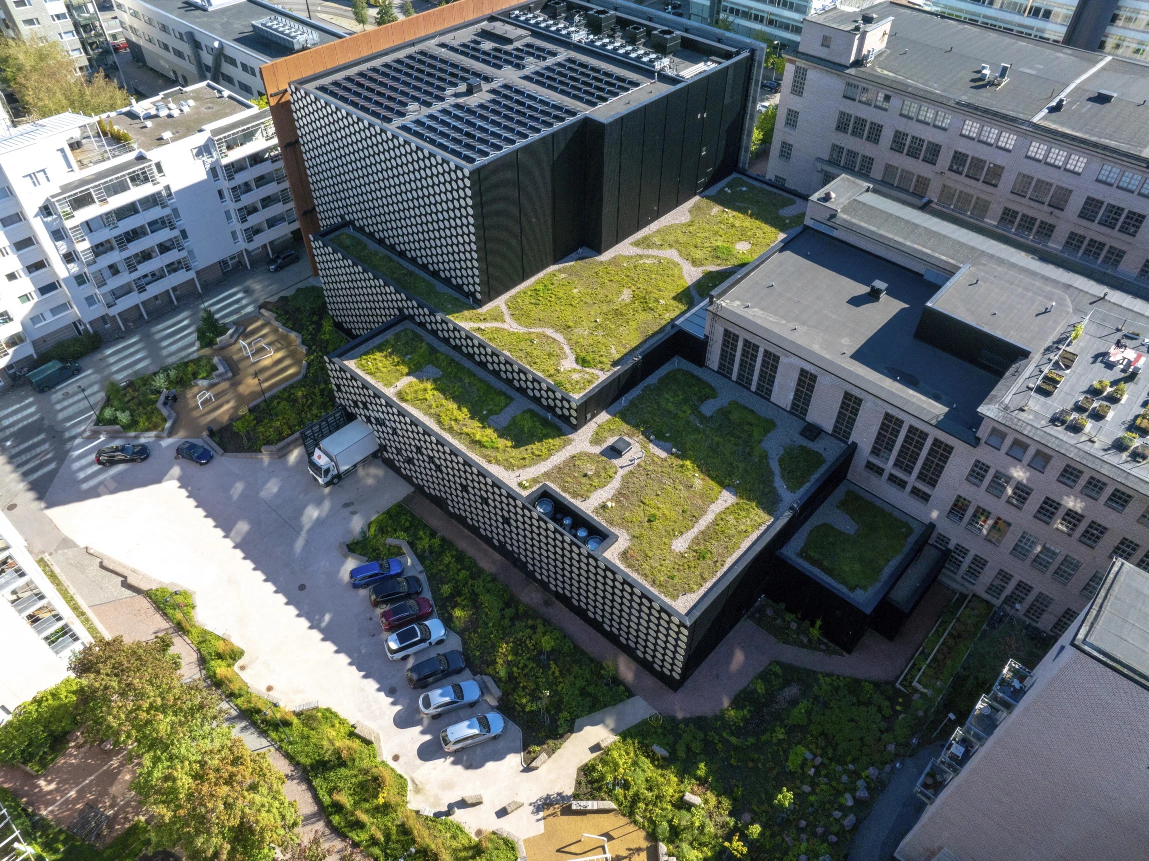 Aerial view of a modern building complex with green roofs, surrounded by parked cars and other buildings.