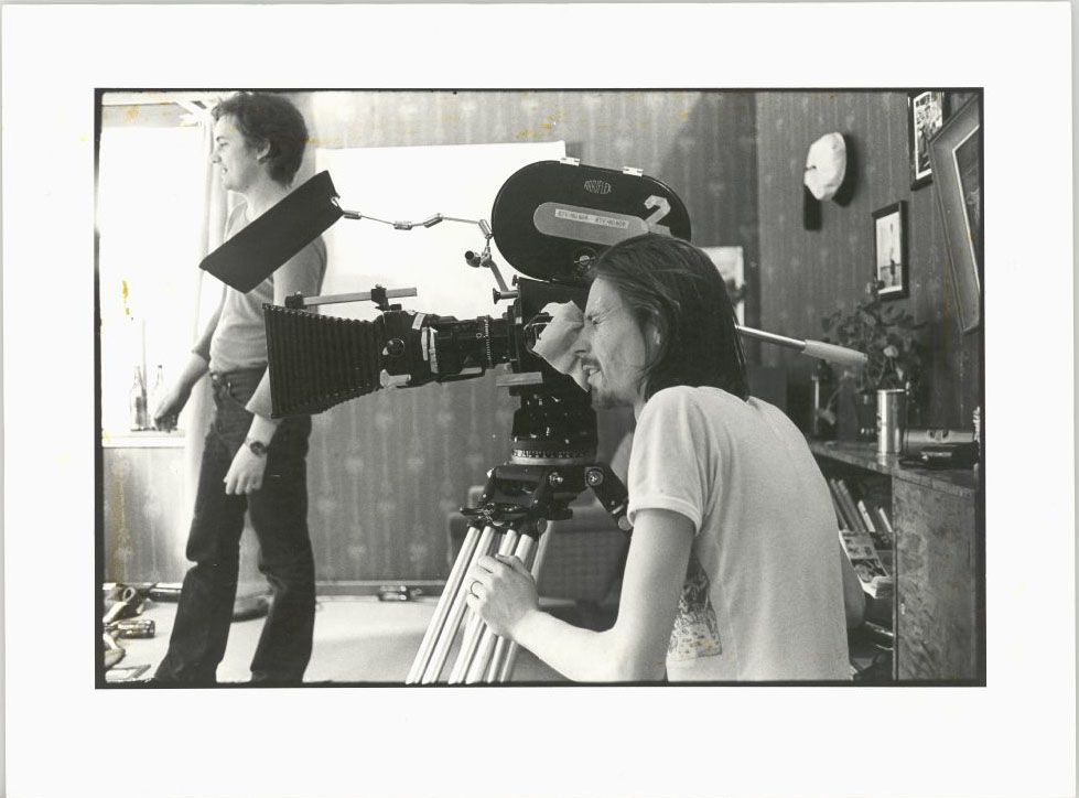 A person operating a vintage film camera on a tripod indoors, with another person standing in the background.