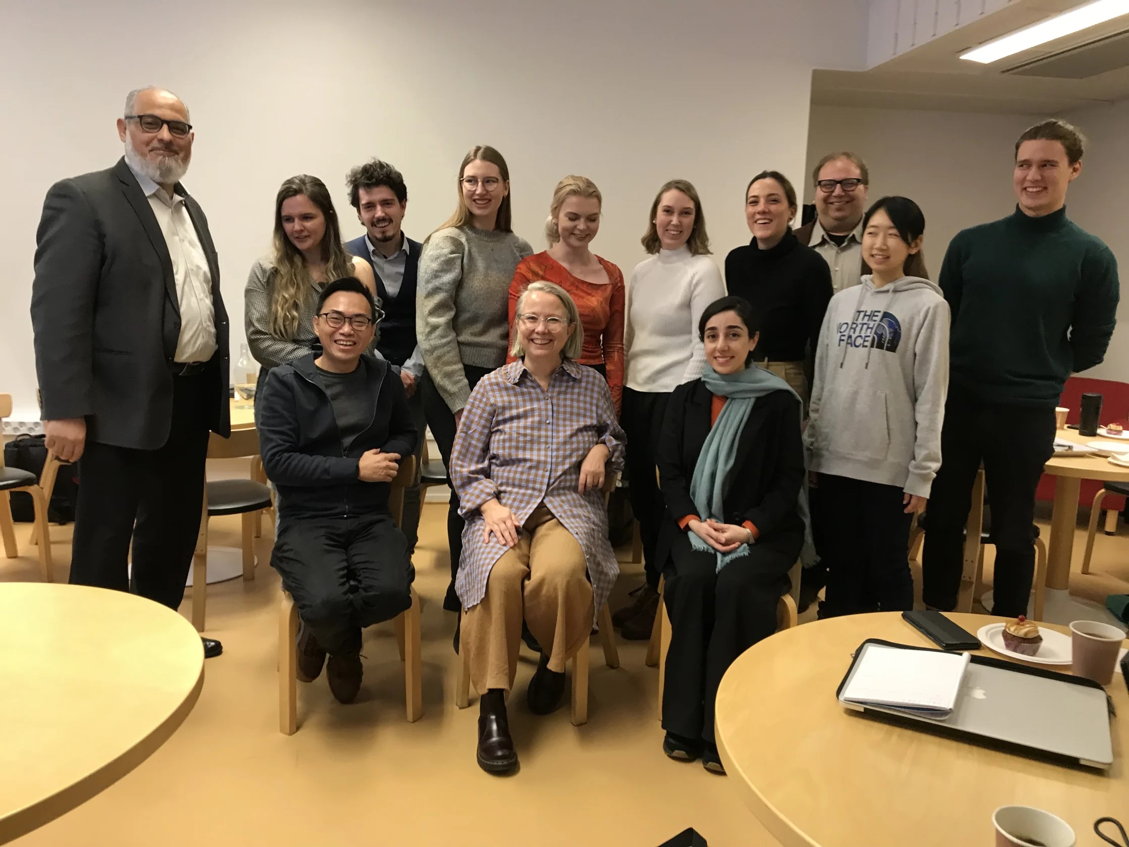 A group of people posing indoors, with one person seated in the centre. Tables and chairs are visible in the background.