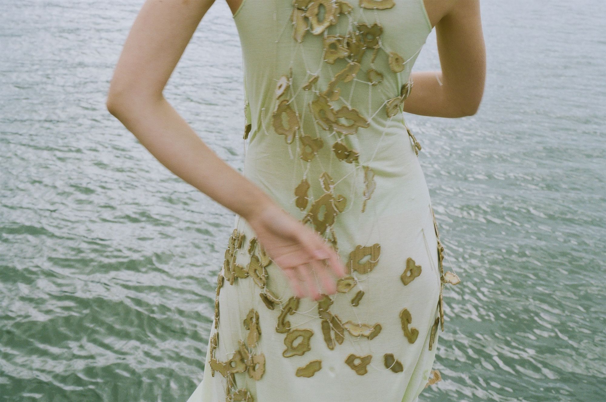 A person in a light green dress with floral patterns stands near a body of water, facing away.