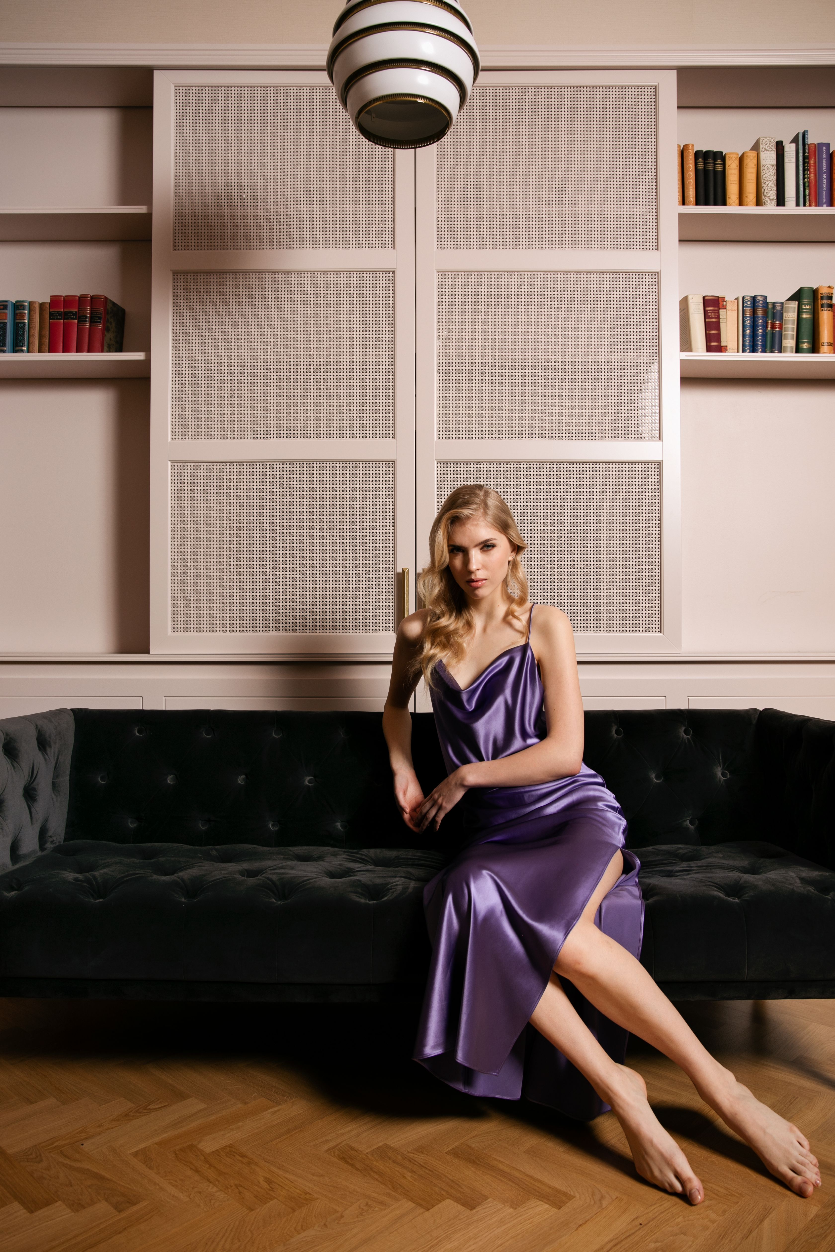 A woman in a purple dress sits on a dark sofa in a room with wooden flooring and bookshelves.