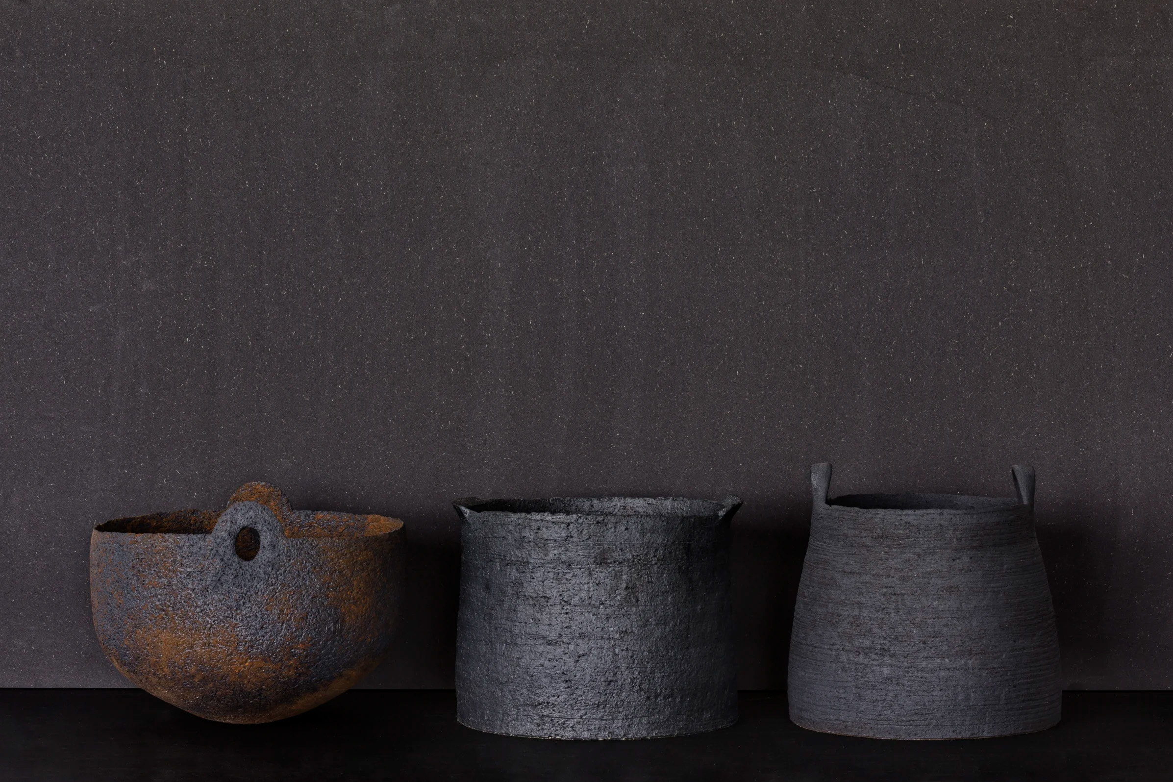 Kolme teksturoitua kulhoa: yksi ruosteisen ruskea reiällä, kaksi tummanharmaata kahvoilla, tummaa taustaa vasten.
