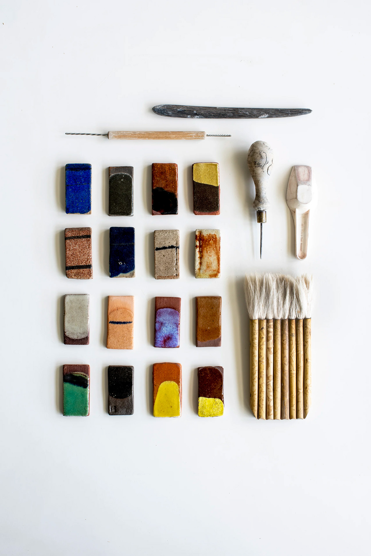 Sarja värikkäitä keraamisia laattoja, työkaluja ja siveltimiä valkoisella pinnalla.