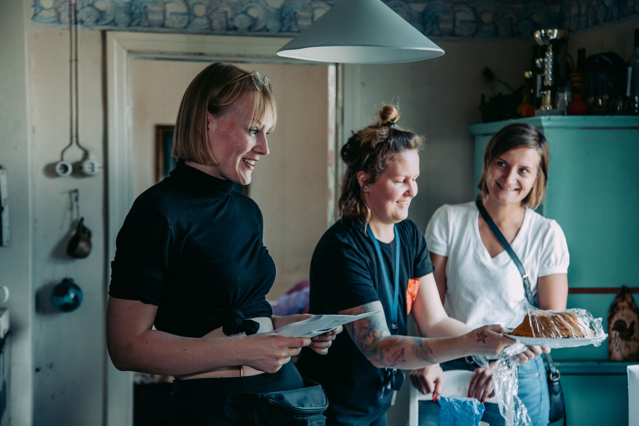 Kolme ihmistä keittiössä, yksi pitää kakkua. Keittiössä on vintage-ilme, turkoosi kaappi ja roikkuvia välineitä.