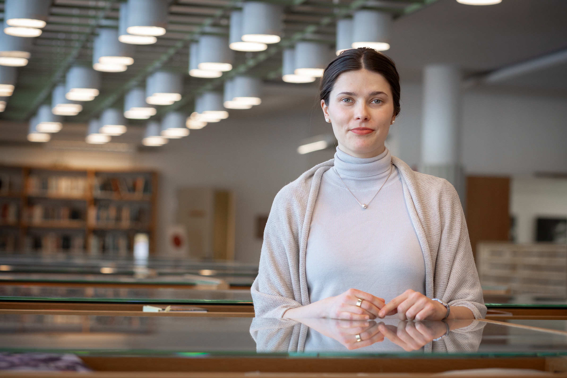 A person stands in a library, wearing a light grey turtleneck and shawl. Bookshelves and ceiling lights are visible in the background.
