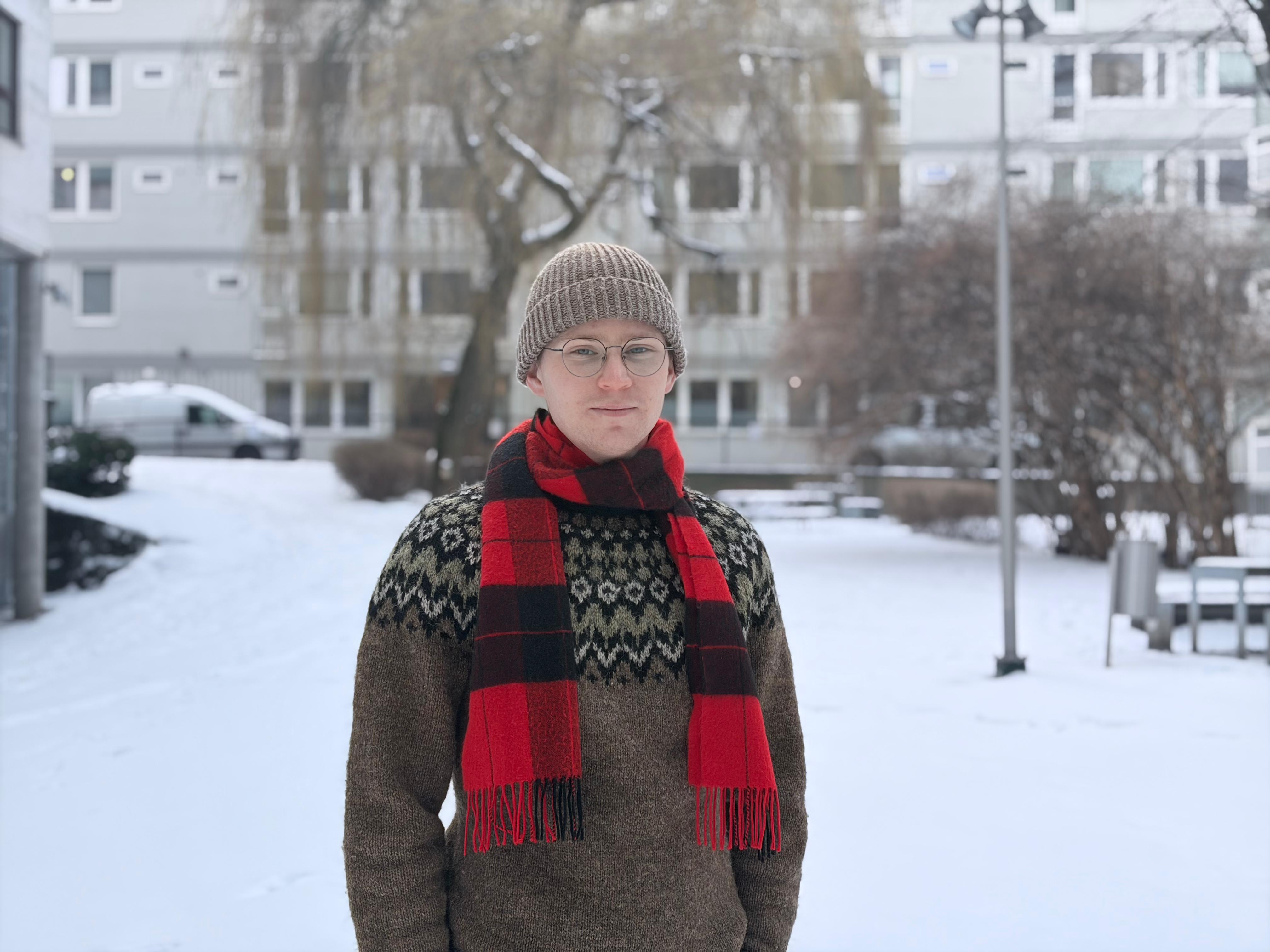 Person in a knitted hat and red scarf standing in a snowy courtyard with buildings and trees in the background.