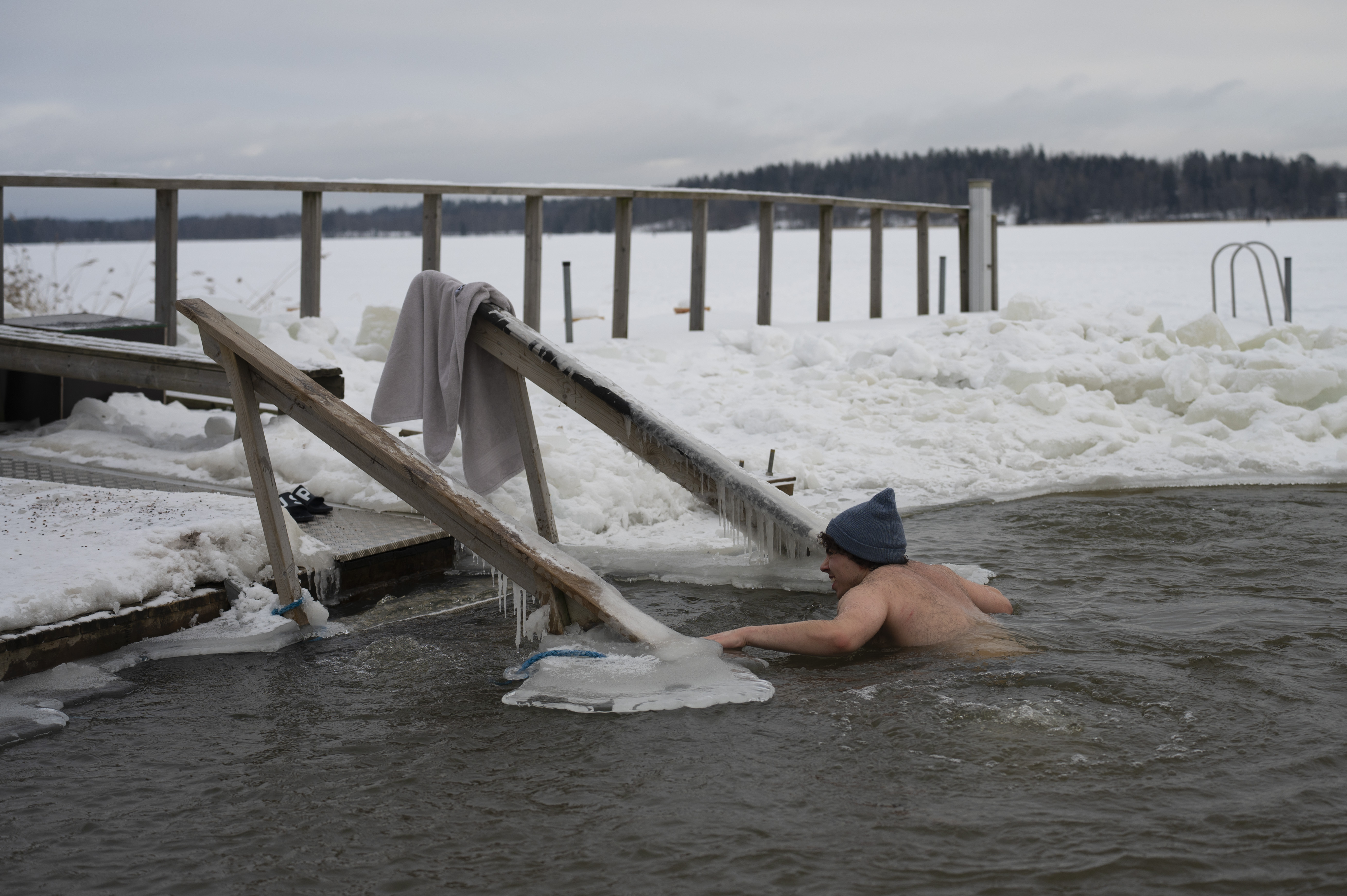A person swims in icy water near a snowy dock with a towel hanging on a wooden railing.