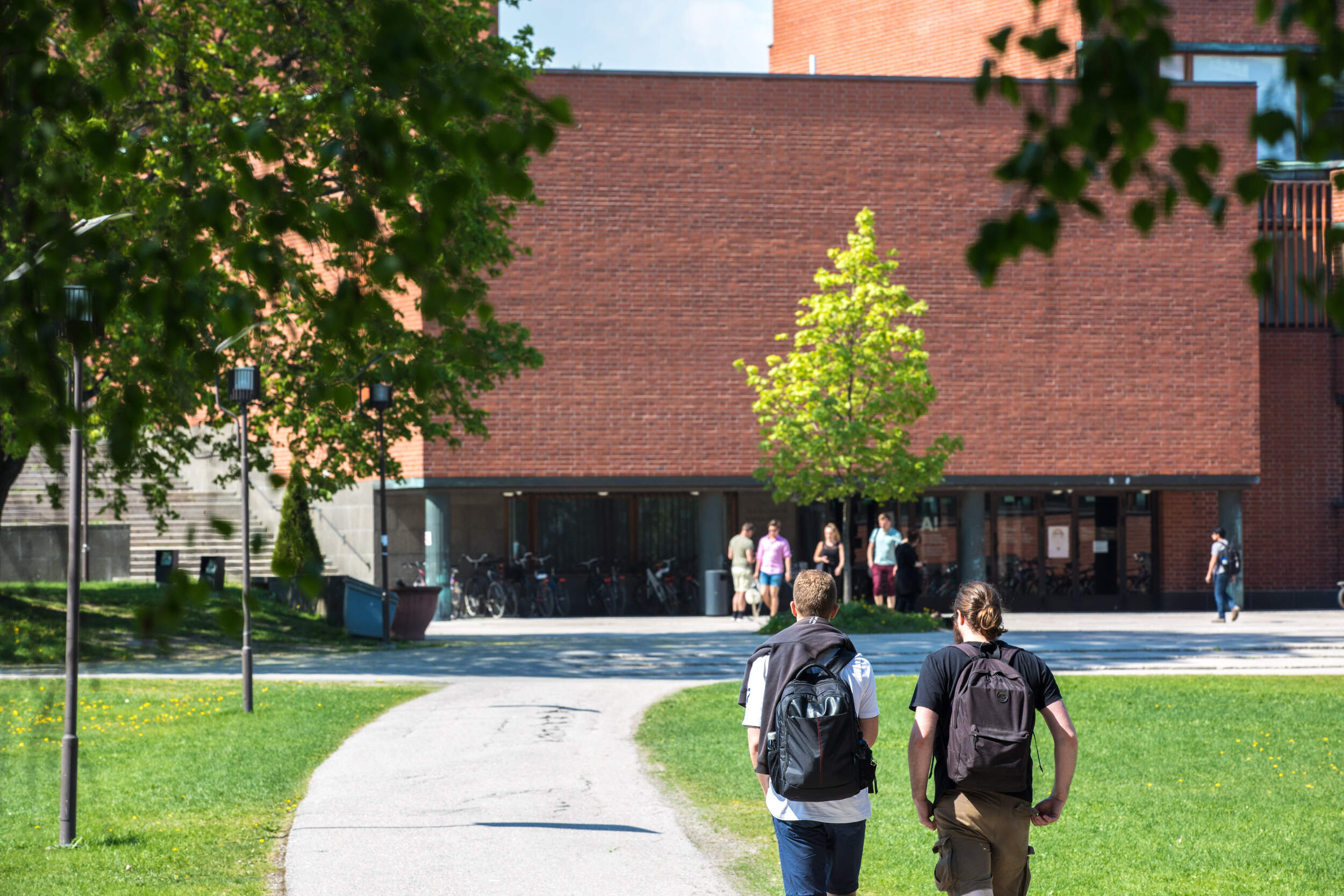Two people with backpacks walk toward a red brick building with a tree in front and several people near the entrance.