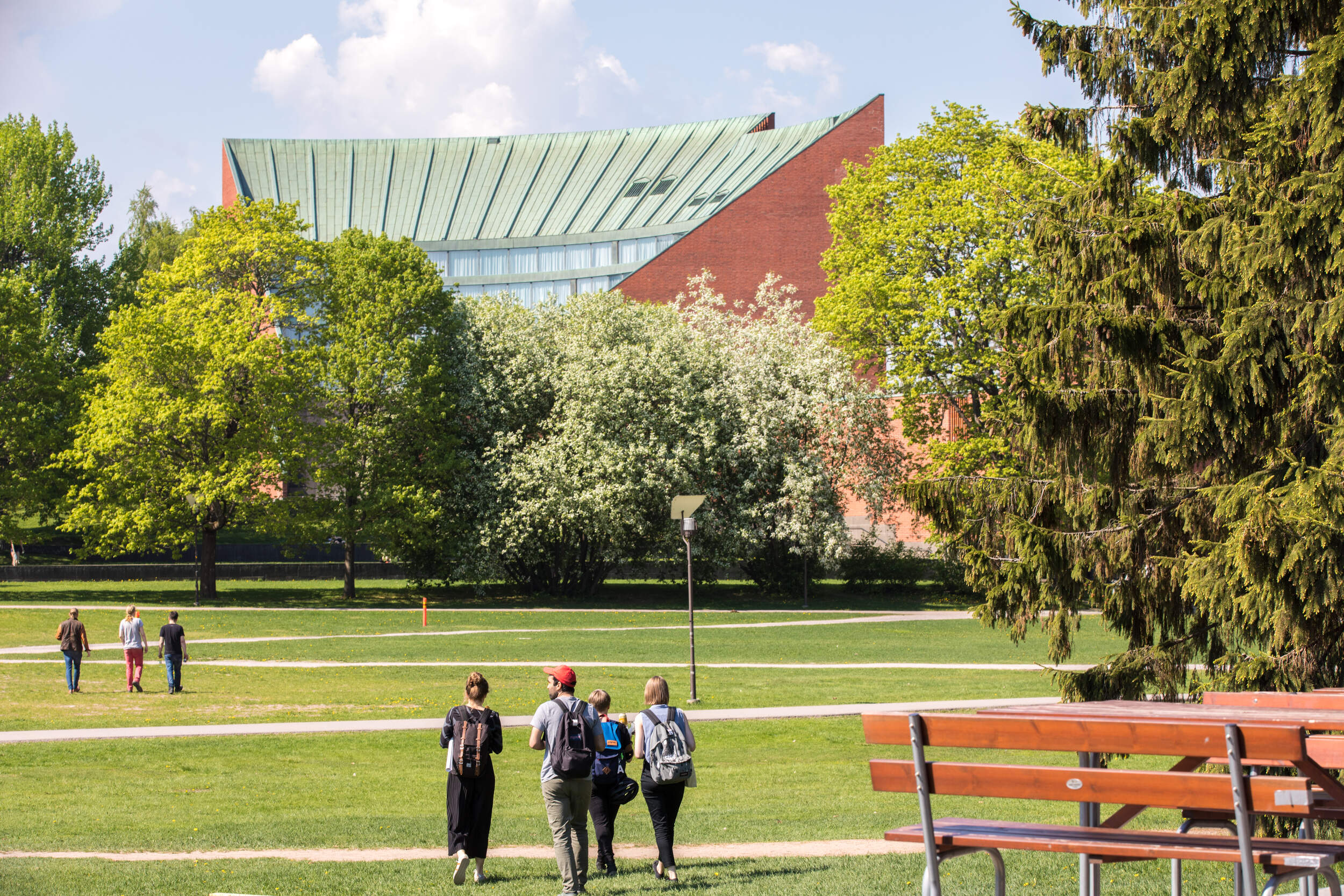 Students walk across a sunny green campus lawn with trees and the Undergraduate Center.