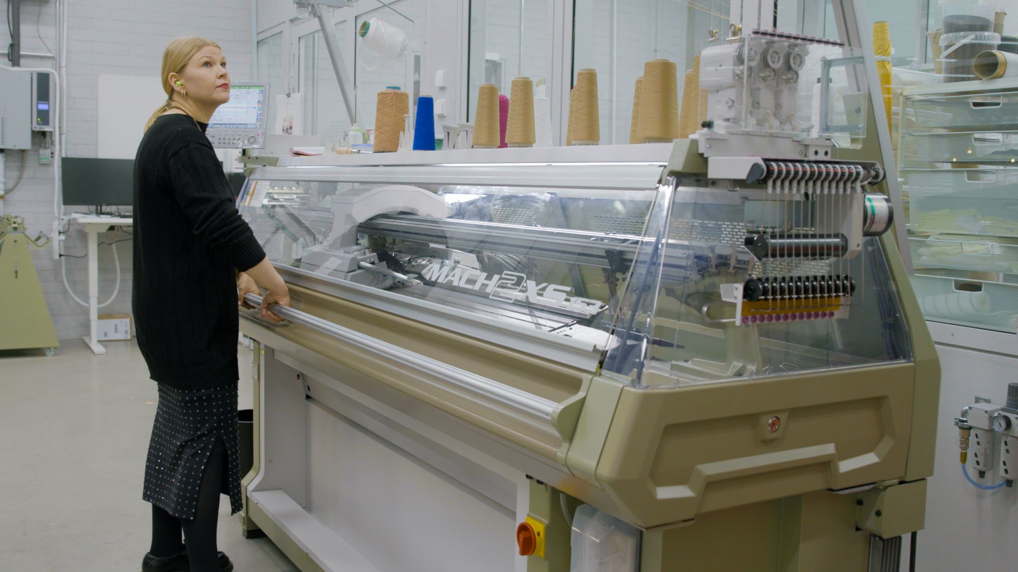 Musta-asuinen henkilö käyttää suurta beigeä neulekoneetta laboratoriossa, kartiolankoja sen päällä.