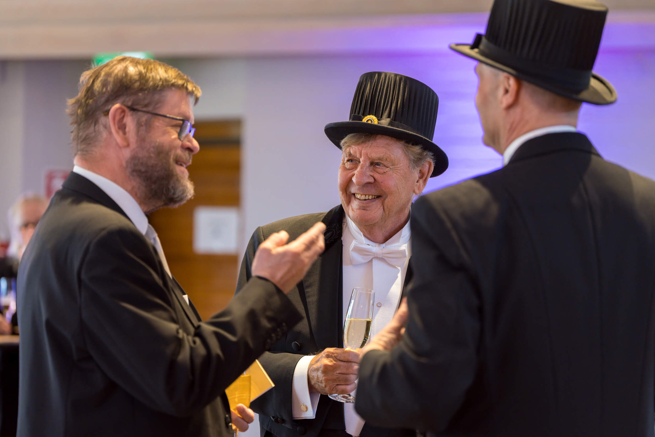 Three formally dressed men in black tailcoats and top hats talk indoors, one holding a champagne glass.