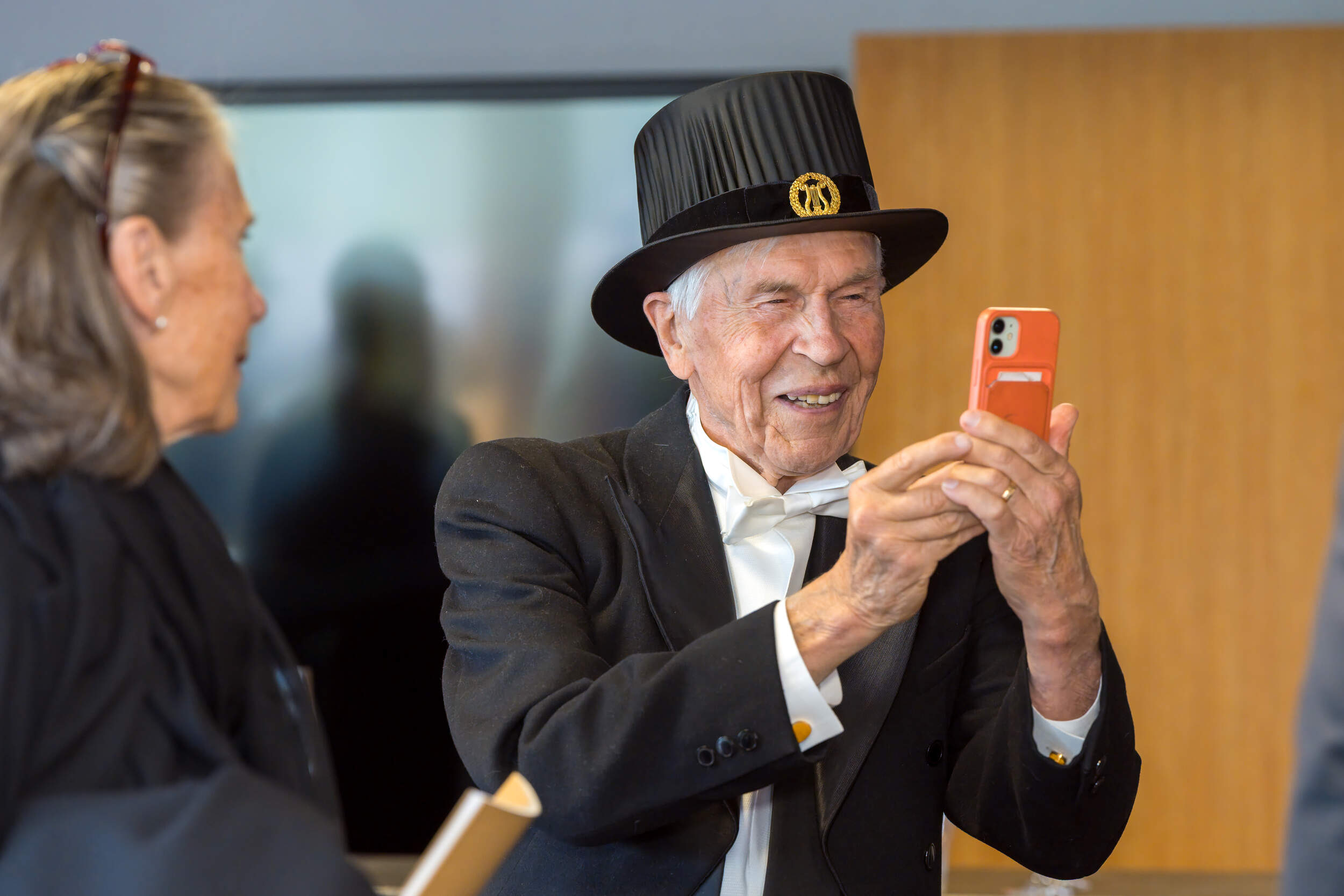 Elderly person in formal black suit and top hat takes a photo with an orange smartphone indoors