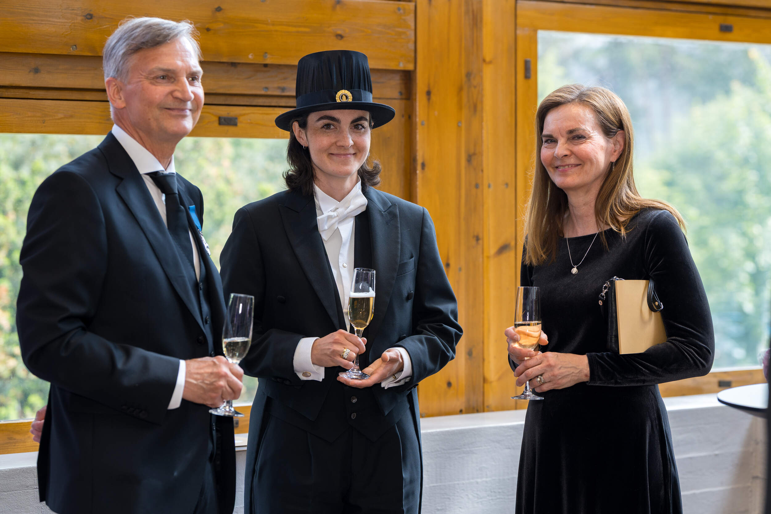 Three formally dressed people at a reception, holding drinks by large wooden-framed windows
