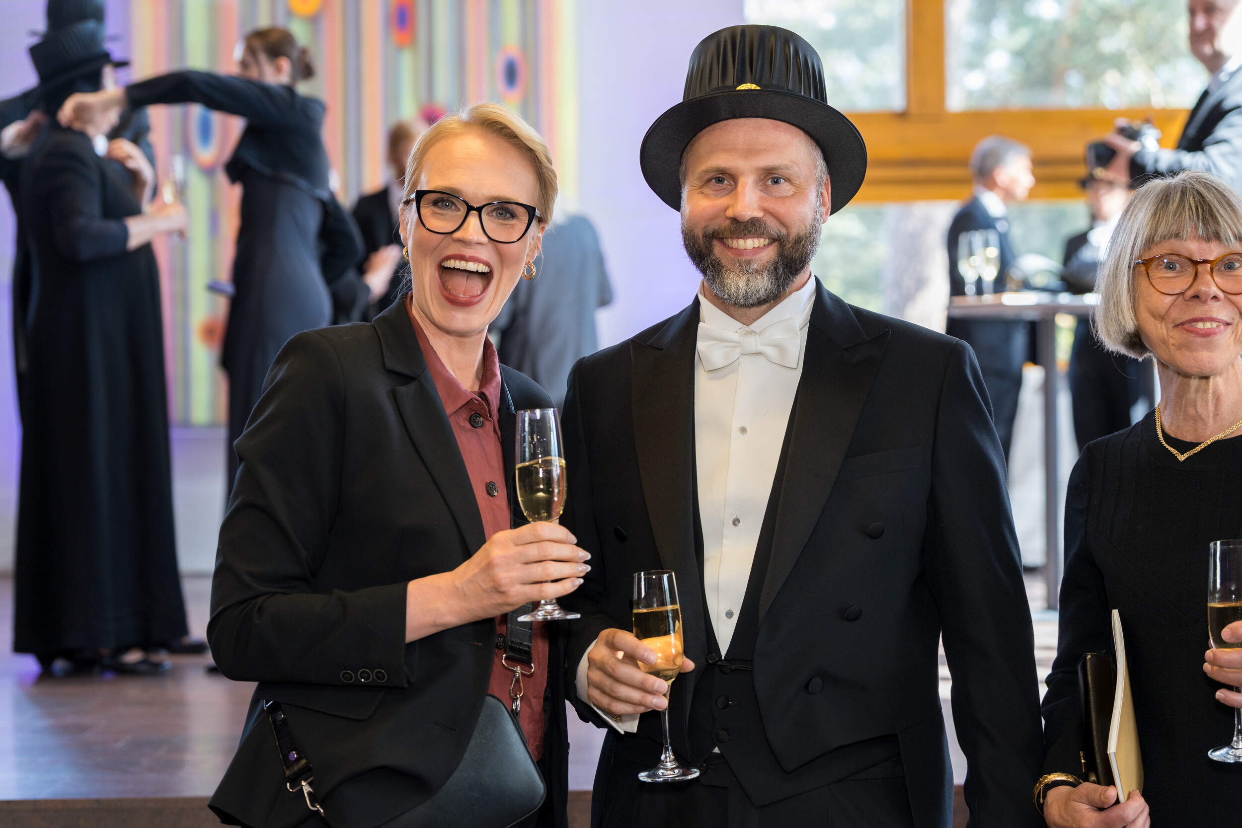 People in formal black outfits holding champagne glasses at an indoor reception with art in the background