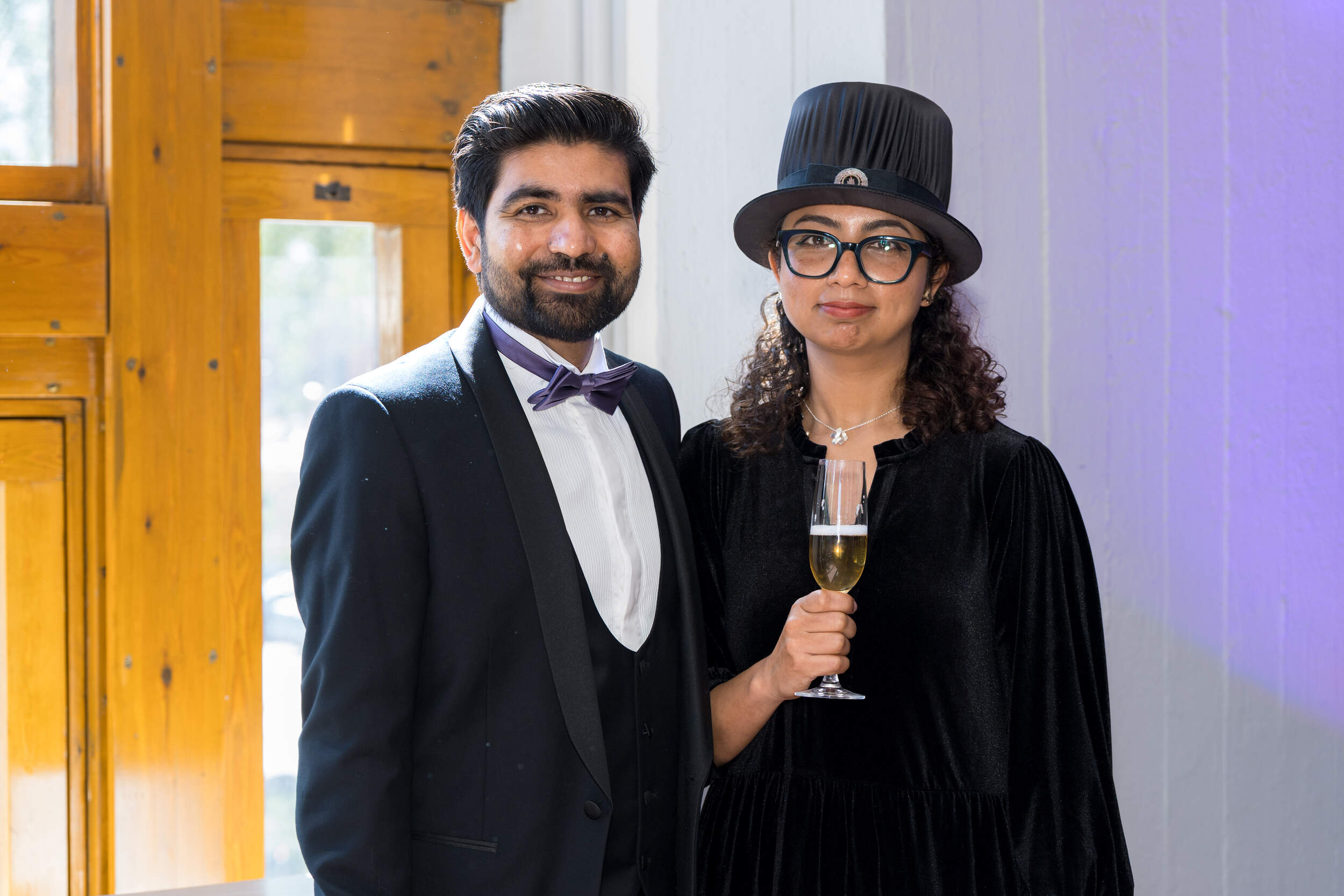 Two formally dressed people indoors; one in a suit, one in a black dress and top hat holding a champagne glass