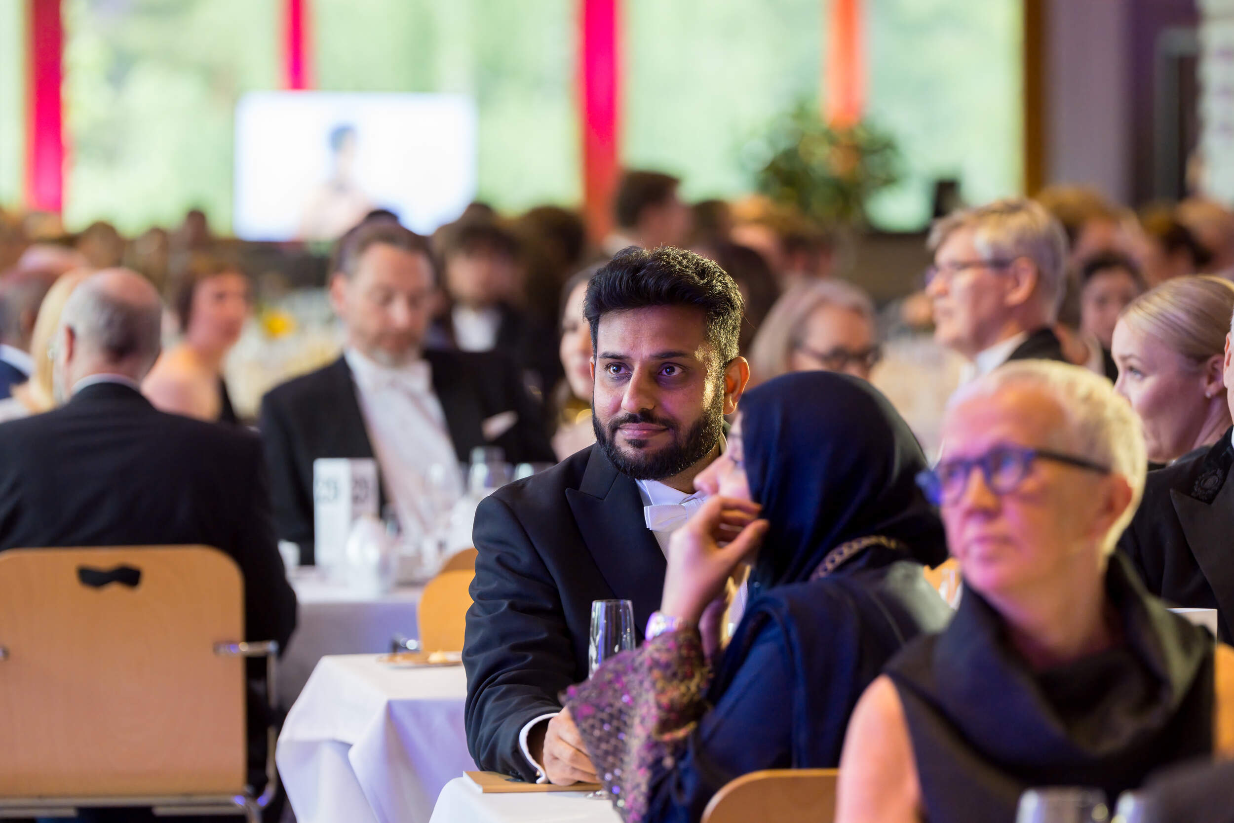 People in formal black and dark clothes seated at round tables during a bright indoor banquet