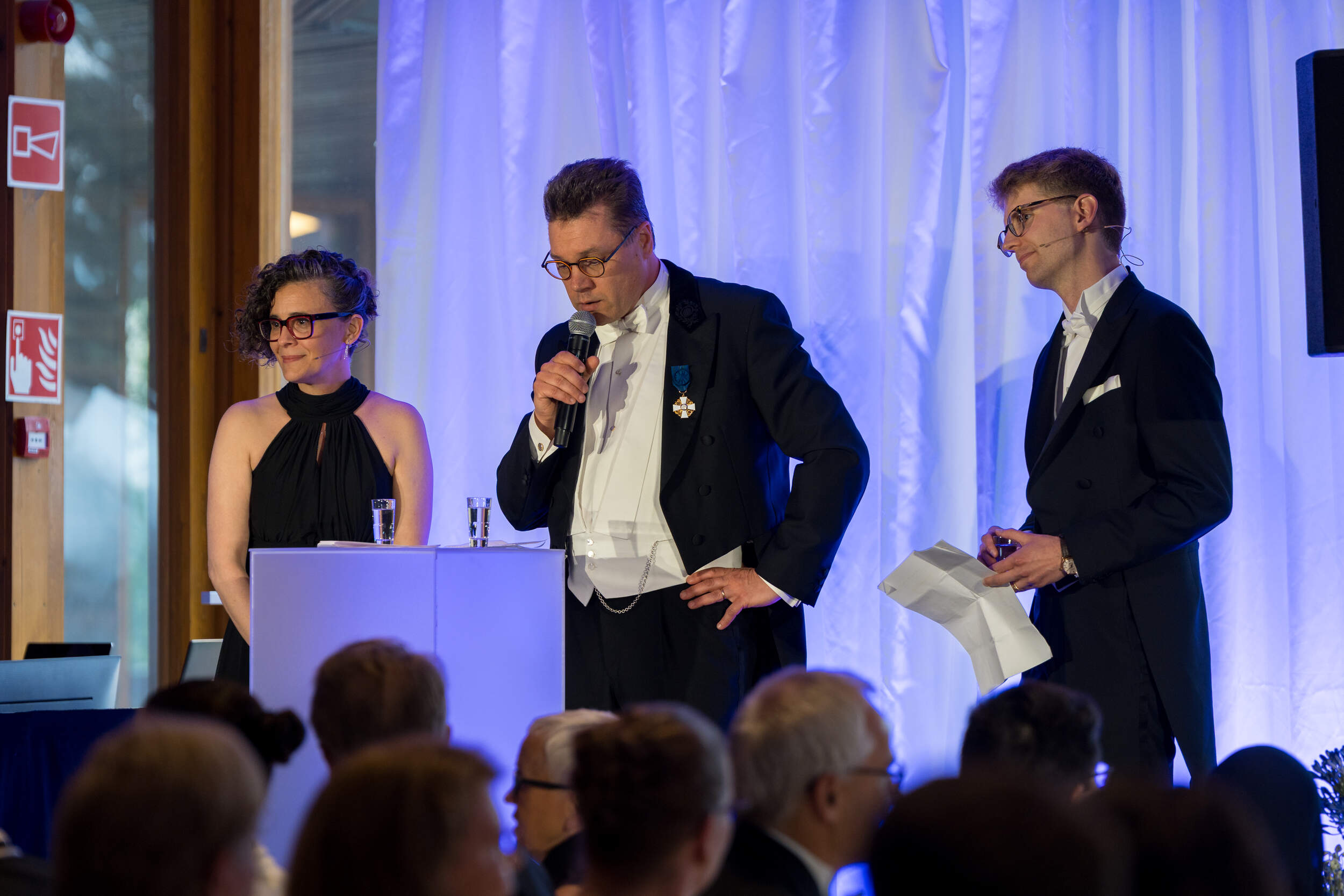 Three speakers in formal black and white evening wear on stage at a blue-lit event