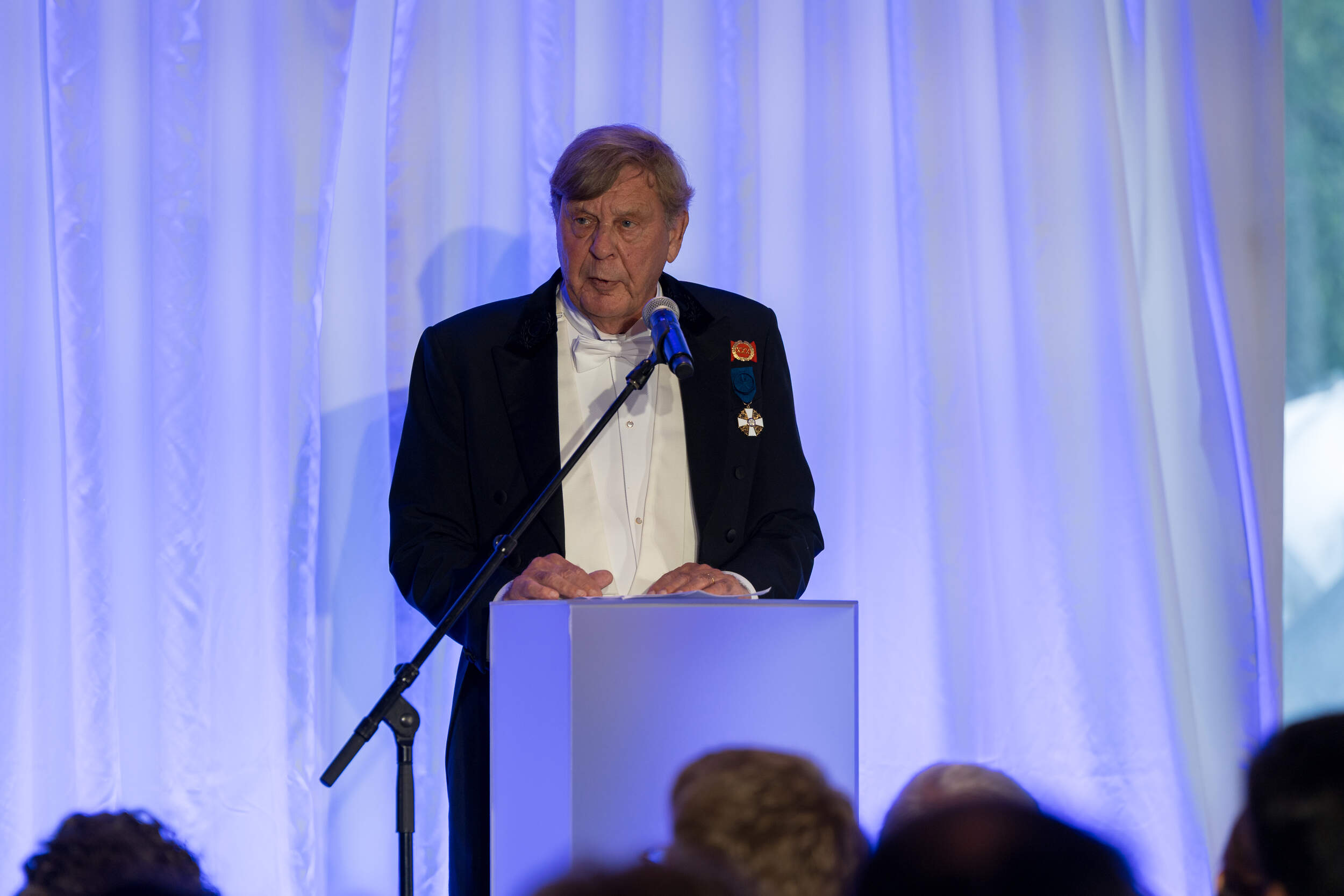 Elderly man in a dark suit with medals speaks at a podium under blue stage lights