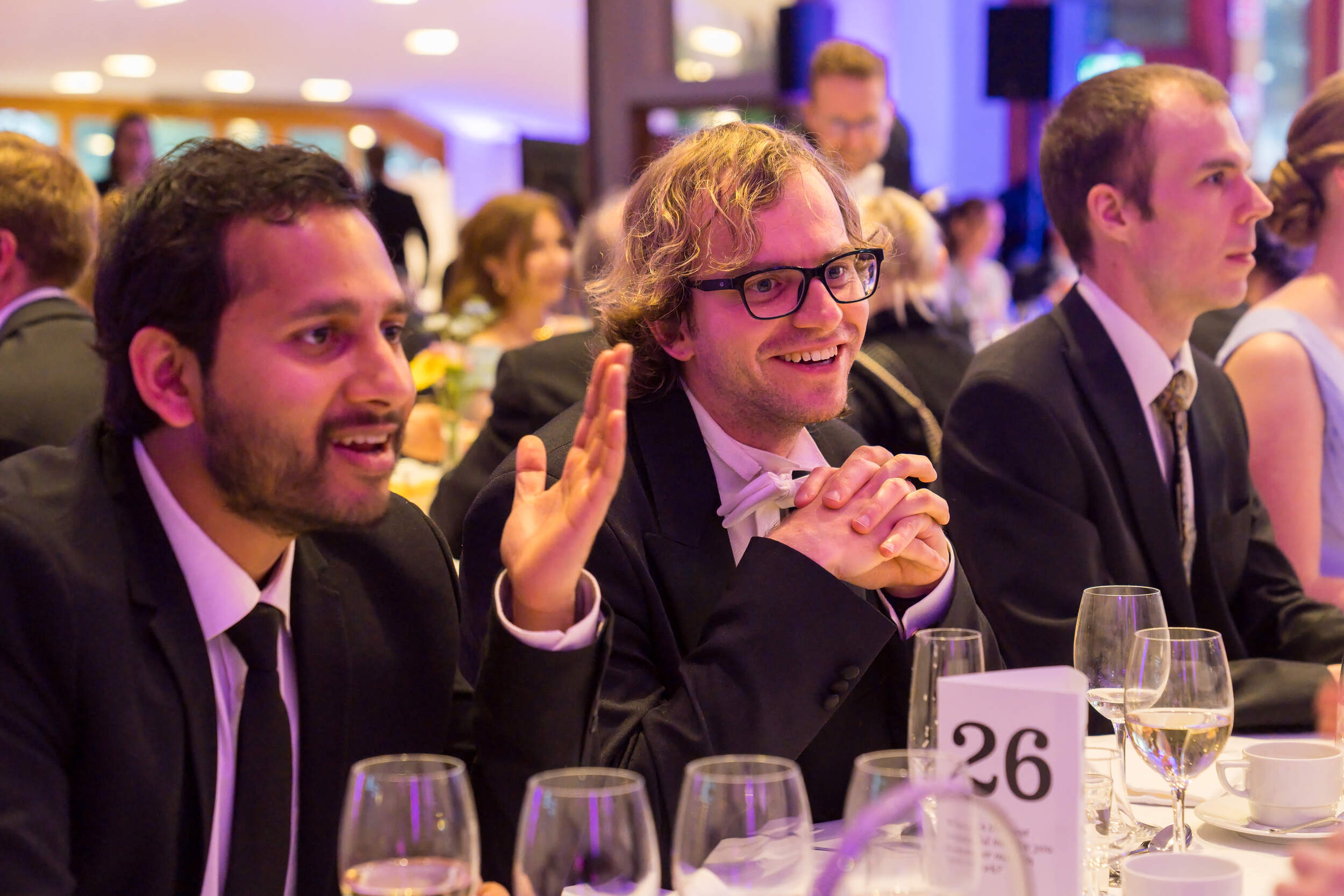 Guests in dark suits sit at a formal dinner table with glasses and a table number card