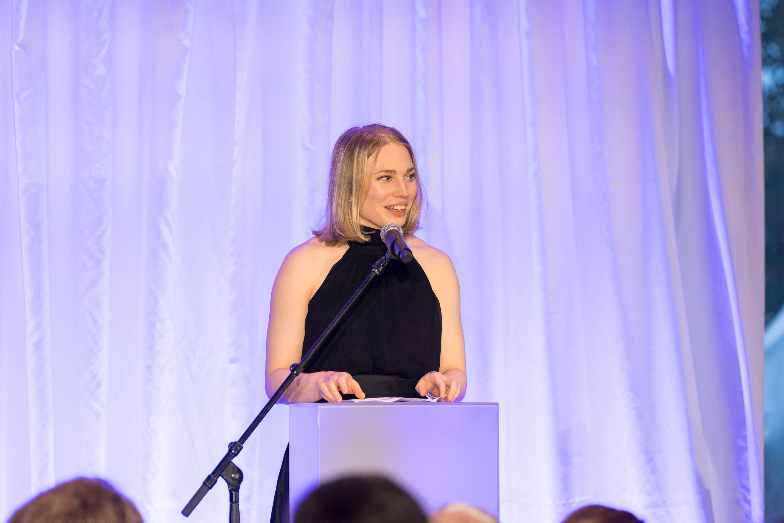 Speaker in a black dress at a podium, addressing an audience against purple-lit curtains