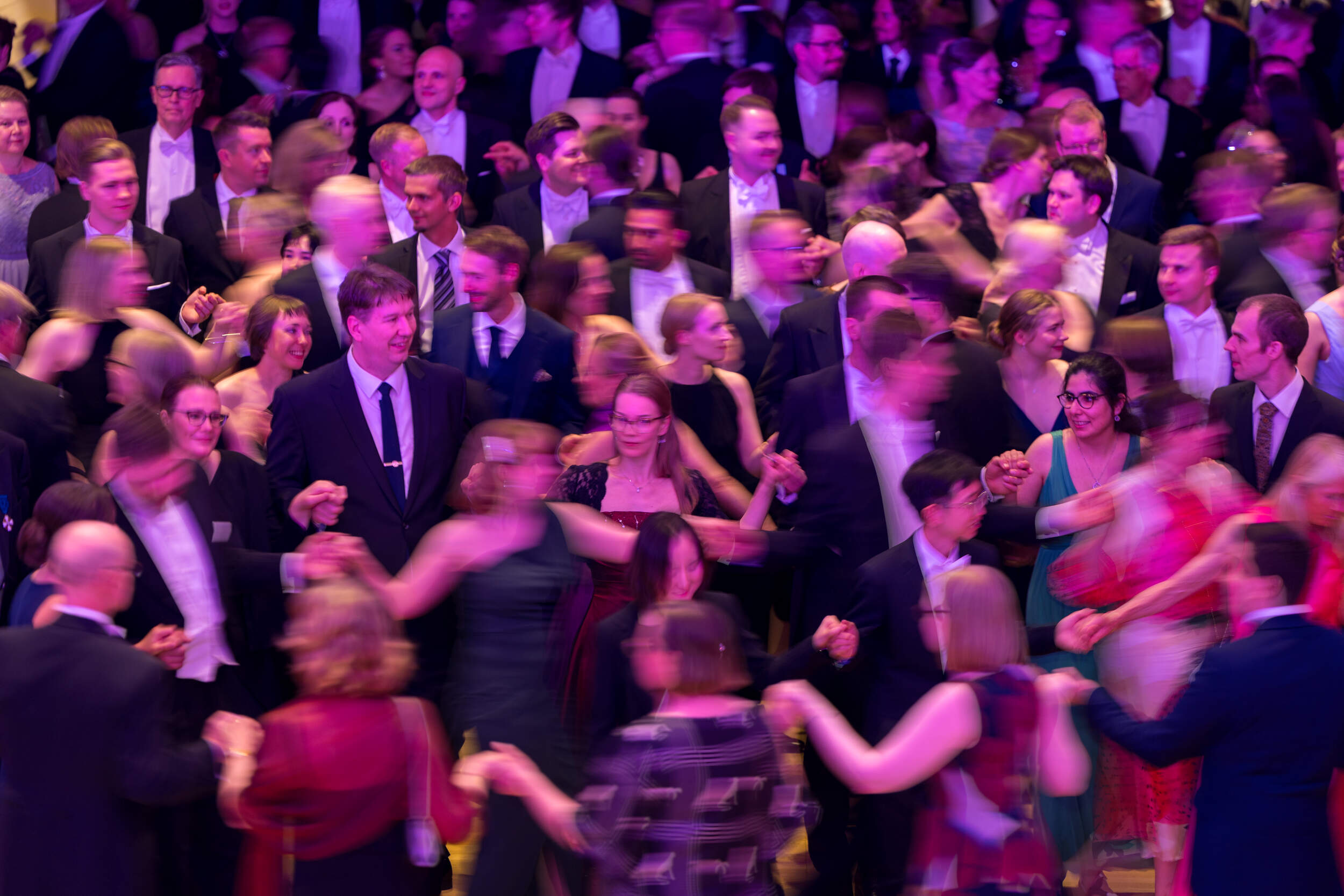 Crowded ballroom with people in formal wear dancing in bright purple and yellow light