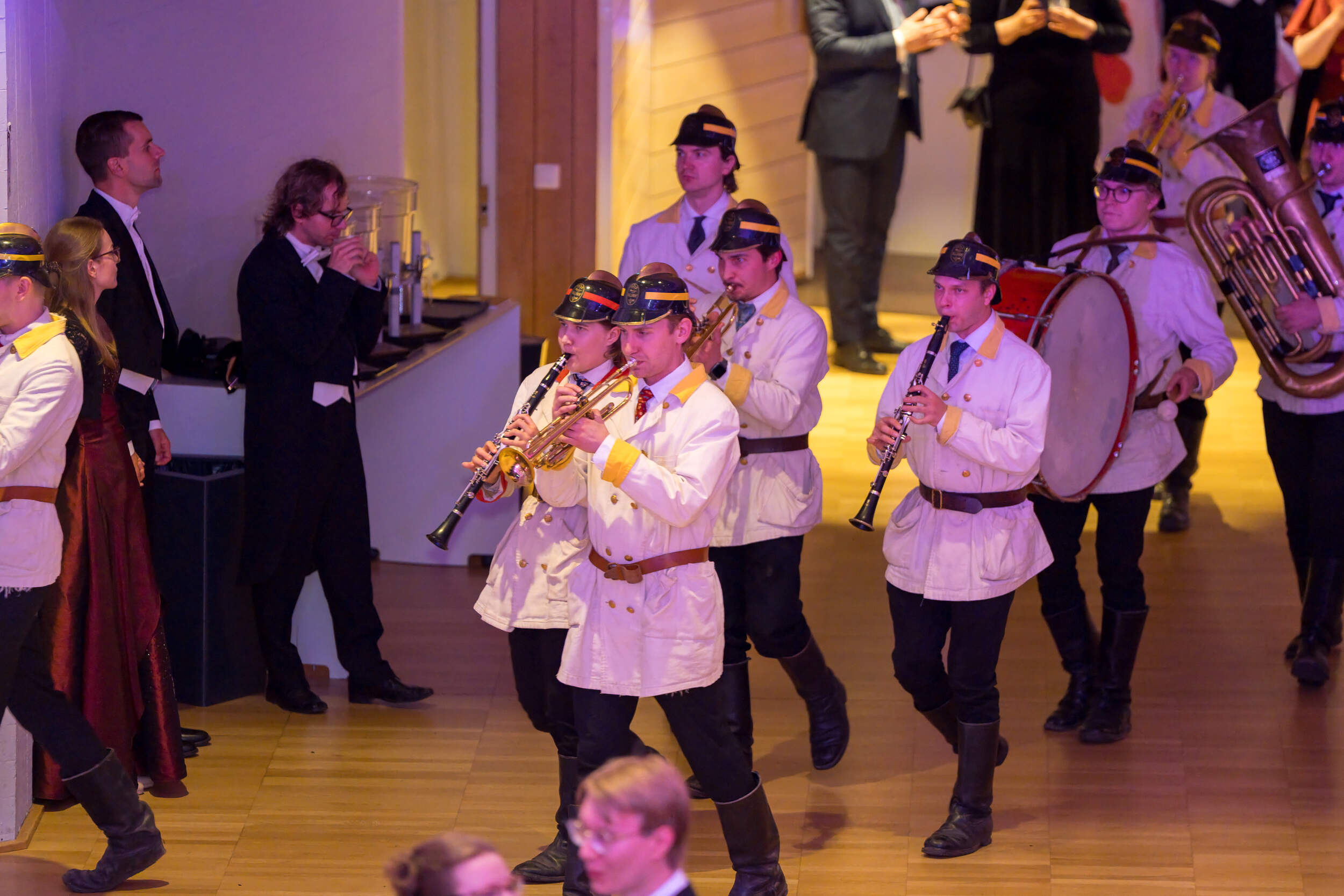 Marching band in white coats and dark hats plays instruments indoors while people in formal wear watch