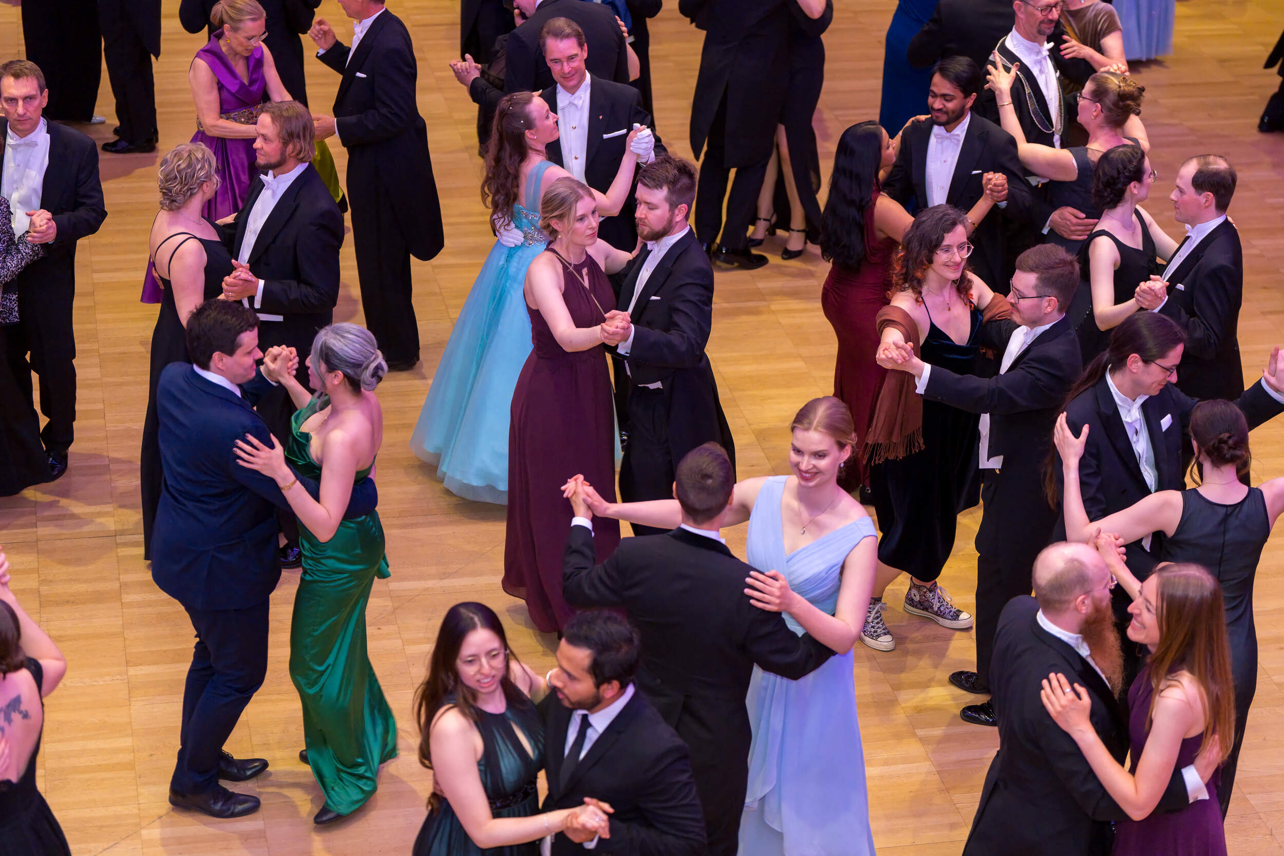 Many couples in formal suits and colourful gowns waltz together on a polished wooden ballroom floor