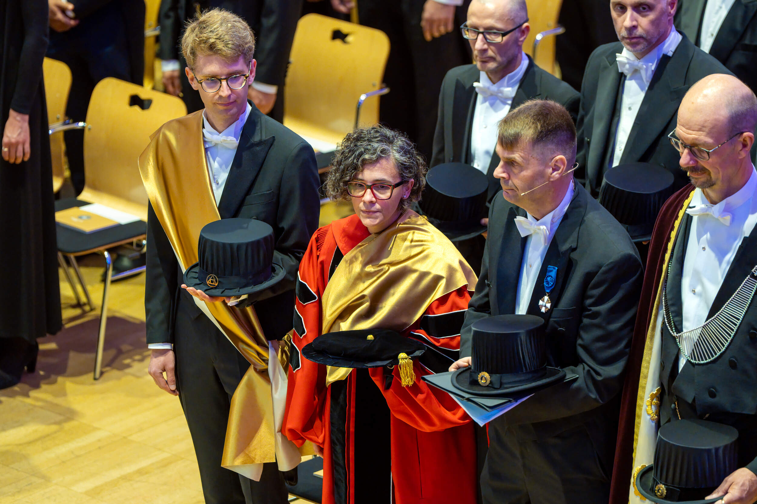 People in formal gowns at a ceremony, holding black academic hats with gold details in a hall