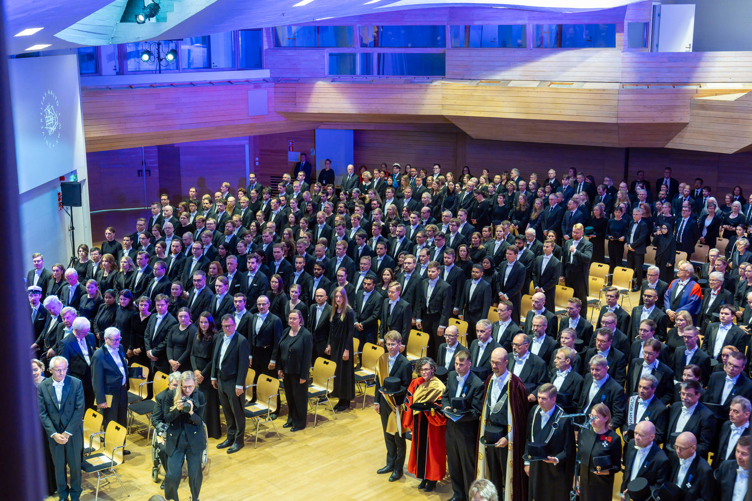 Large formal audience in suits and gowns standing in a modern wood hall with blue lighting