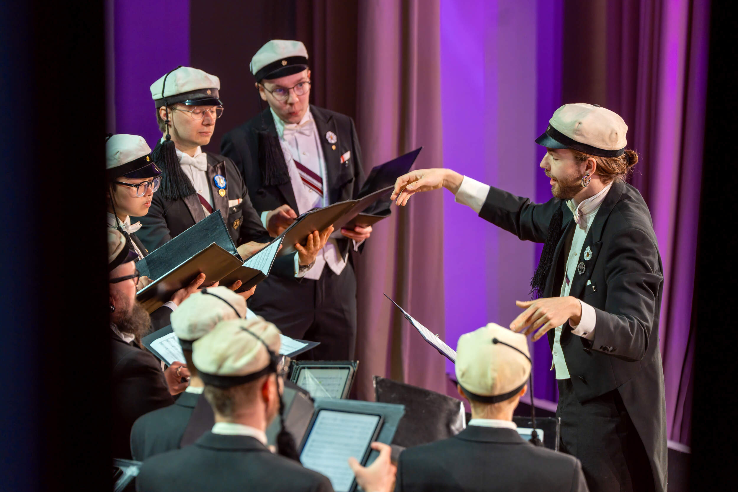 Choir in dark suits and white caps sings on stage as a conductor leads under purple light