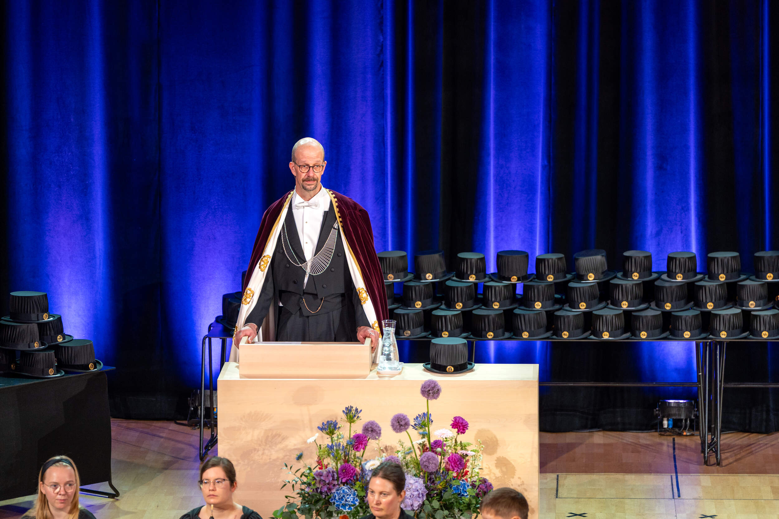 Robed speaker at a podium on stage with doctoral hats and flowers, before blue curtains