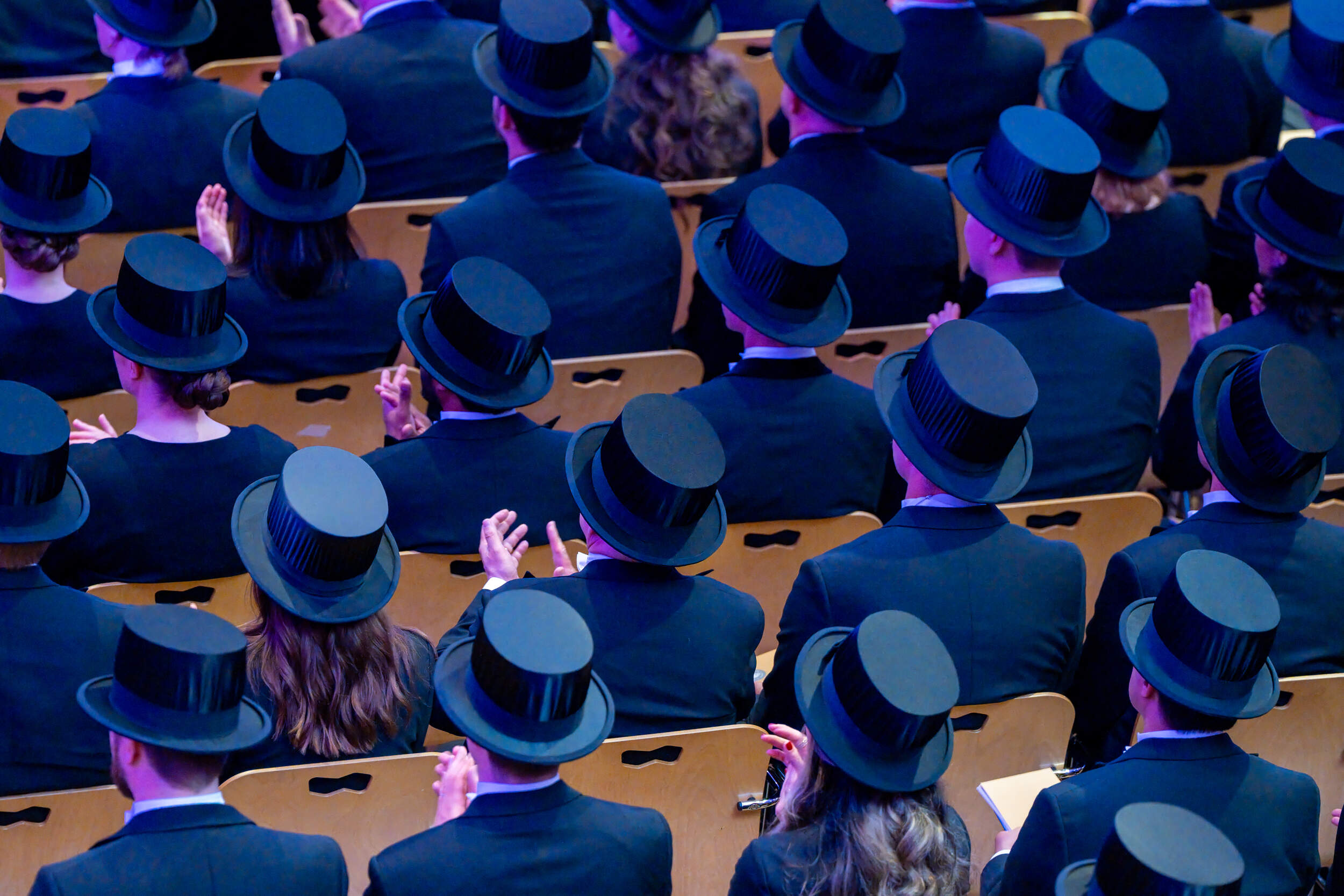 Audience in dark suits and black top hats seated in rows of wooden chairs at a formal ceremony