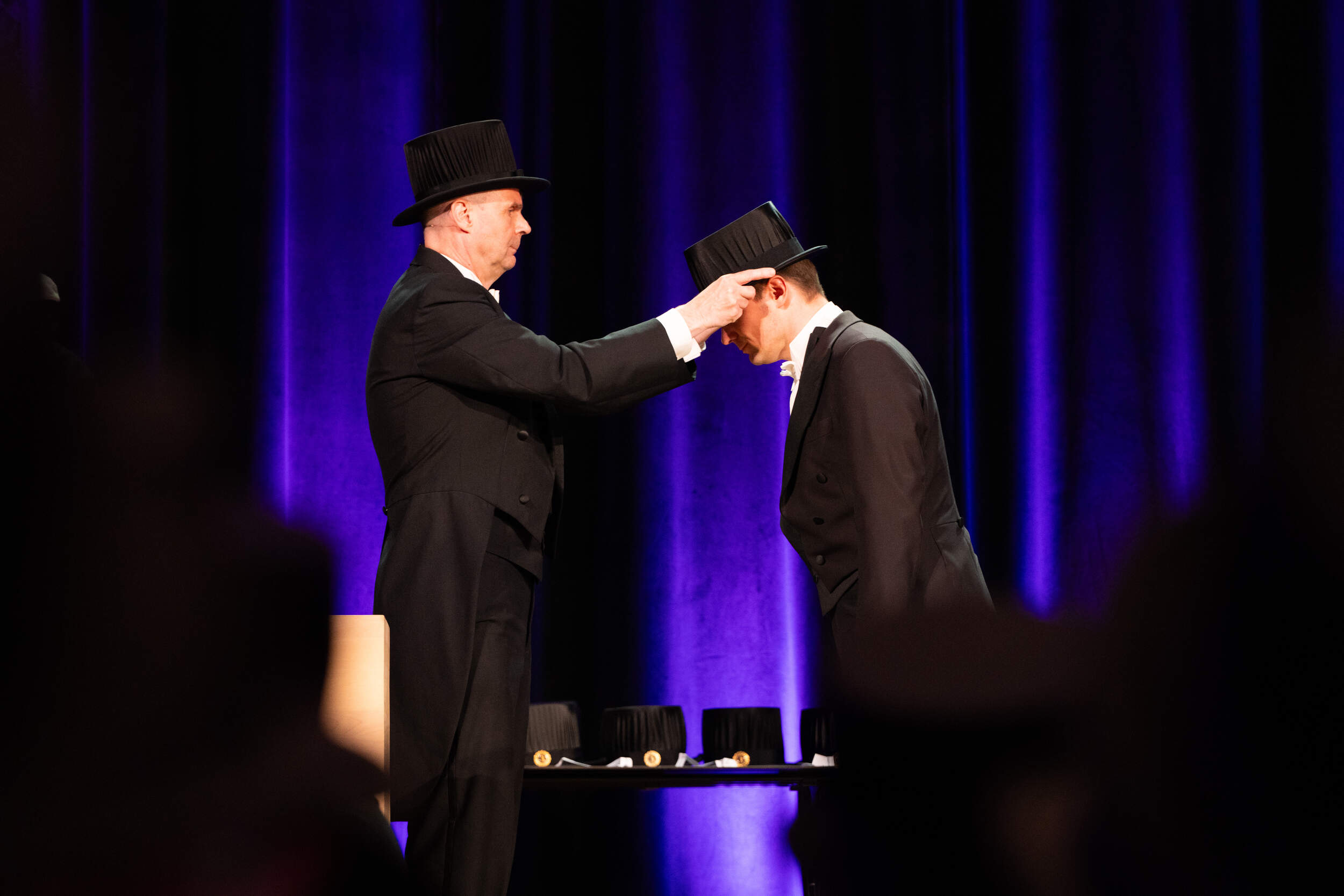 Formal ceremony: person in black tails crowns another with a top hat on a stage lit by purple light.