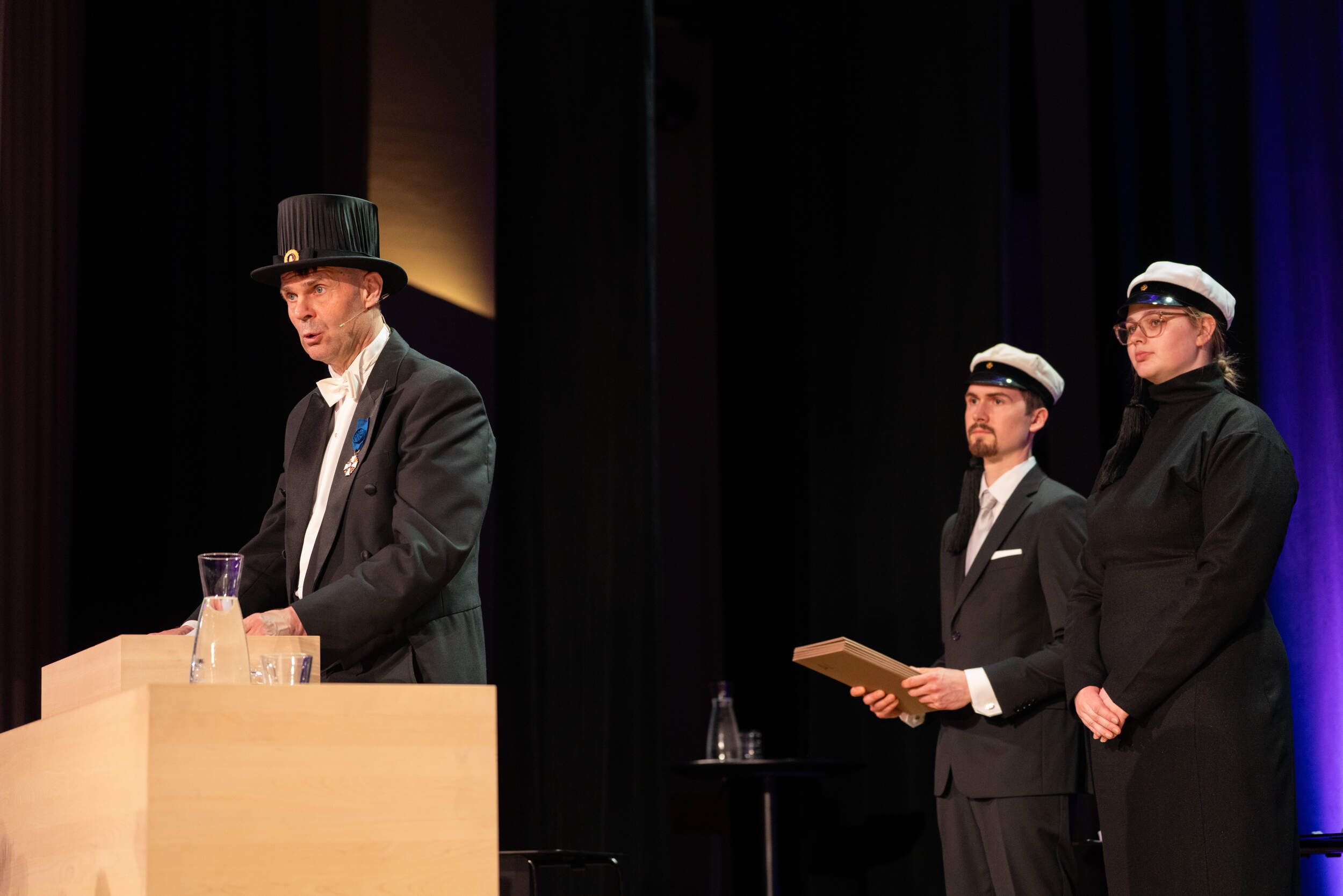 Formal ceremony on stage, three people in dark outfits and white caps, one speaking at a wooden podium